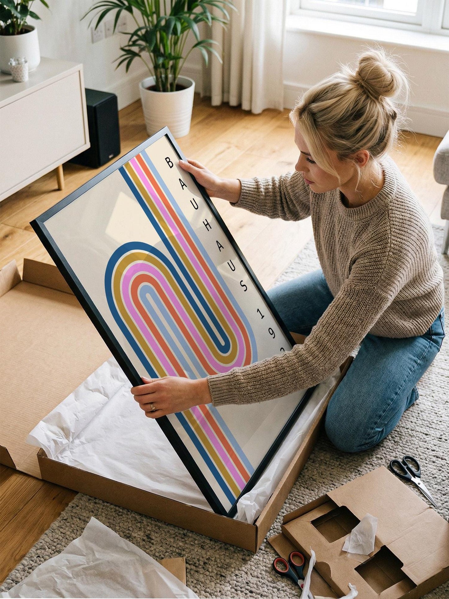 A woman is kneeling on the floor and carefully opening a large cardboard box, which contains a framed piece of art.