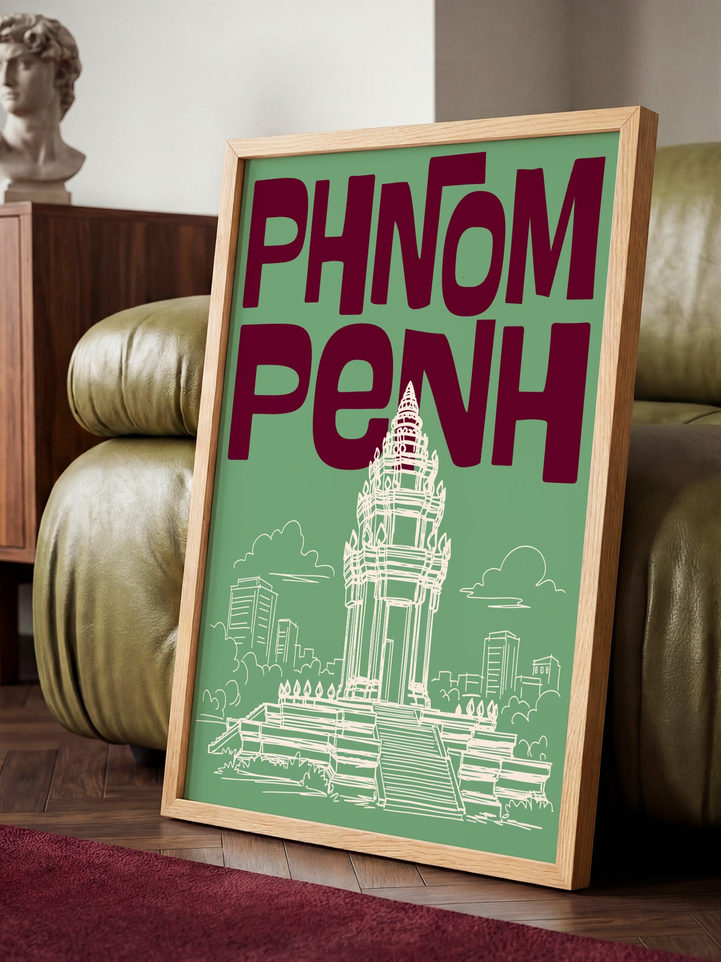 A framed poster with the text "PHNOM PENH" and an illustration of a temple or monument, placed on a wooden floor in a room with a couch and a statue.