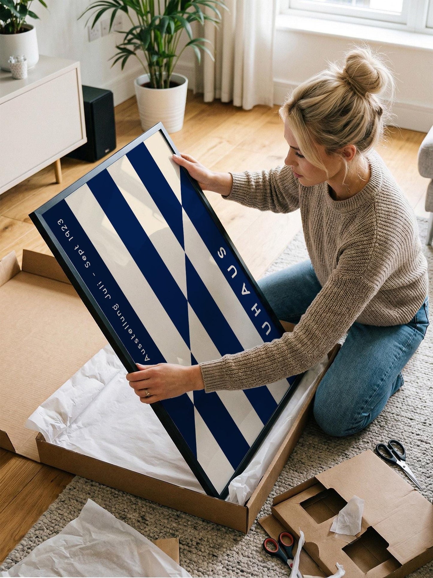 A woman is kneeling on the floor and carefully opening a large cardboard box, which appears to contain a framed artwork.