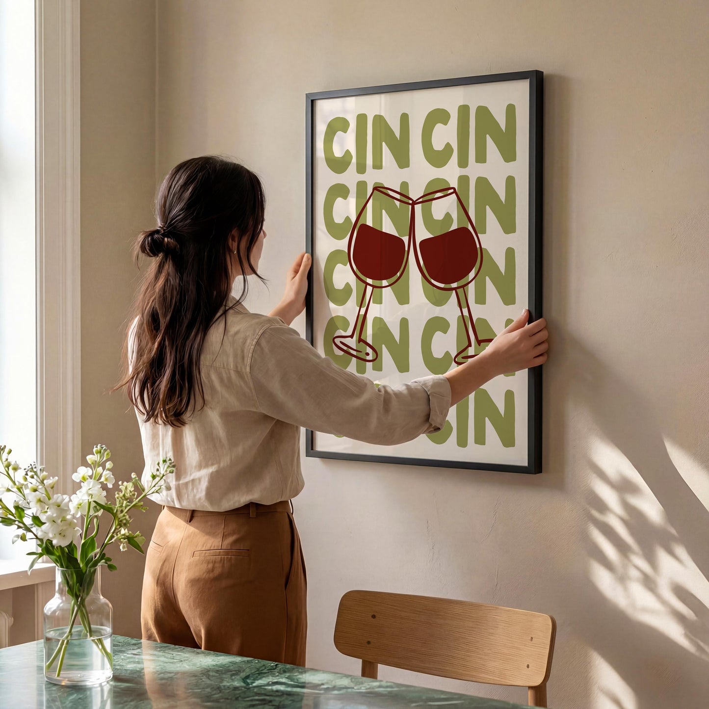 A woman is hanging a framed poster with the word "CIN" on a wall in a room with a dining table and chair visible.