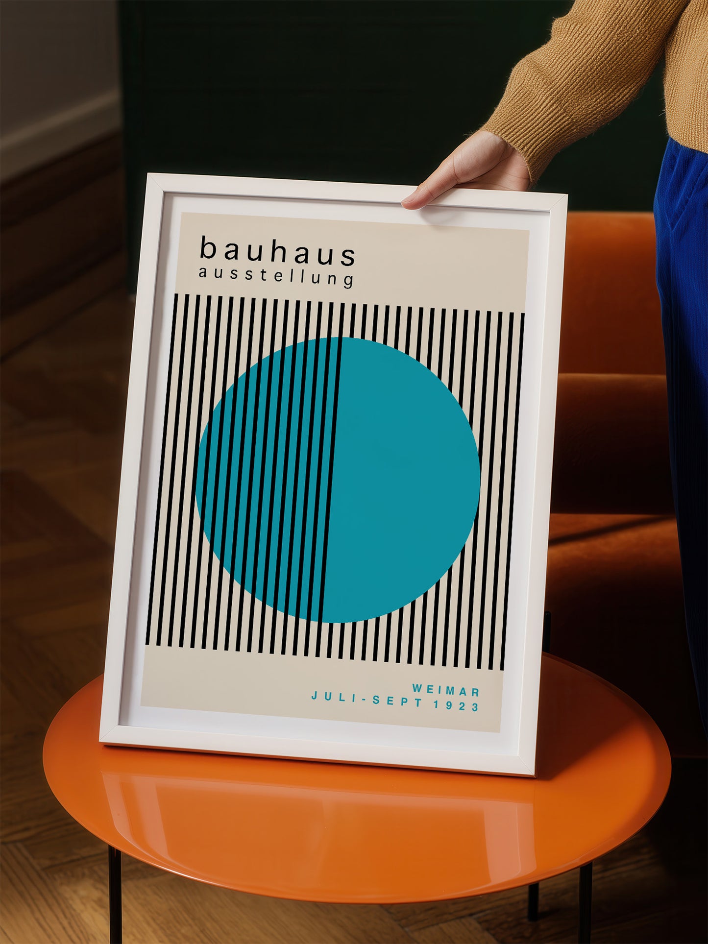 A person is holding a framed poster with a minimalist design featuring a large blue circle and black vertical lines.