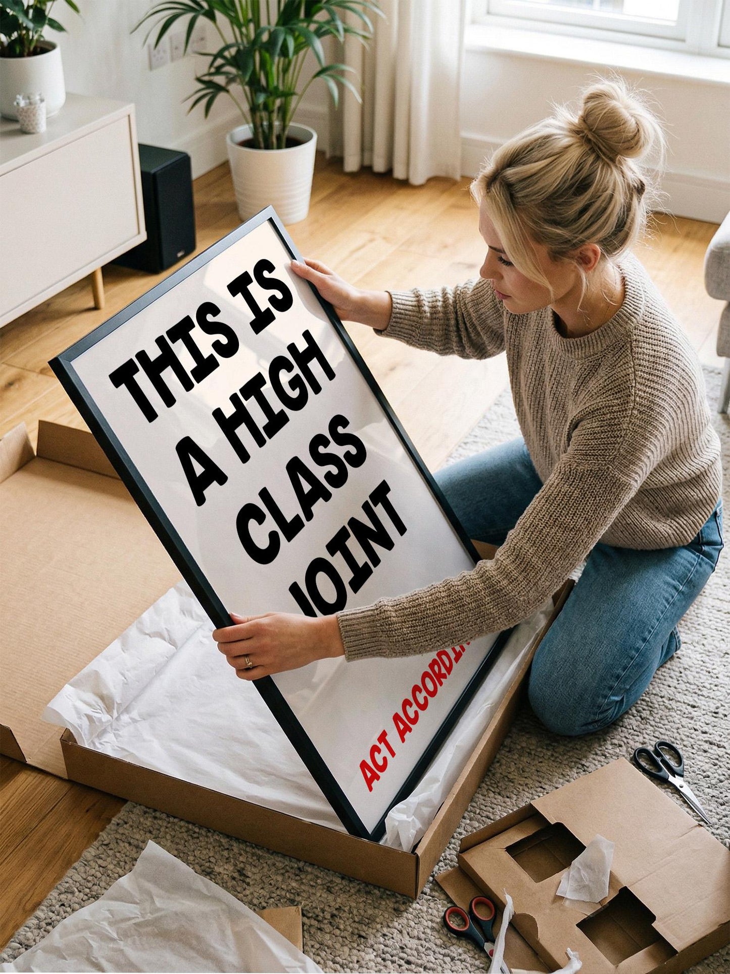 A woman is kneeling on the floor and holding a large framed sign with the text "THIS IS A HIGH CLASS JOINT" on it.