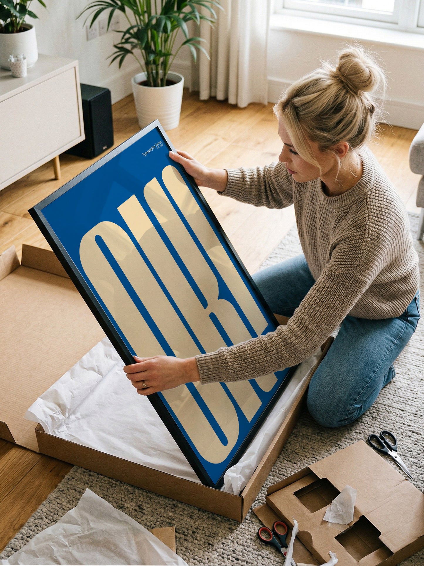 A woman is kneeling on the floor and opening a large cardboard box, which contains a large blue and white poster or artwork.