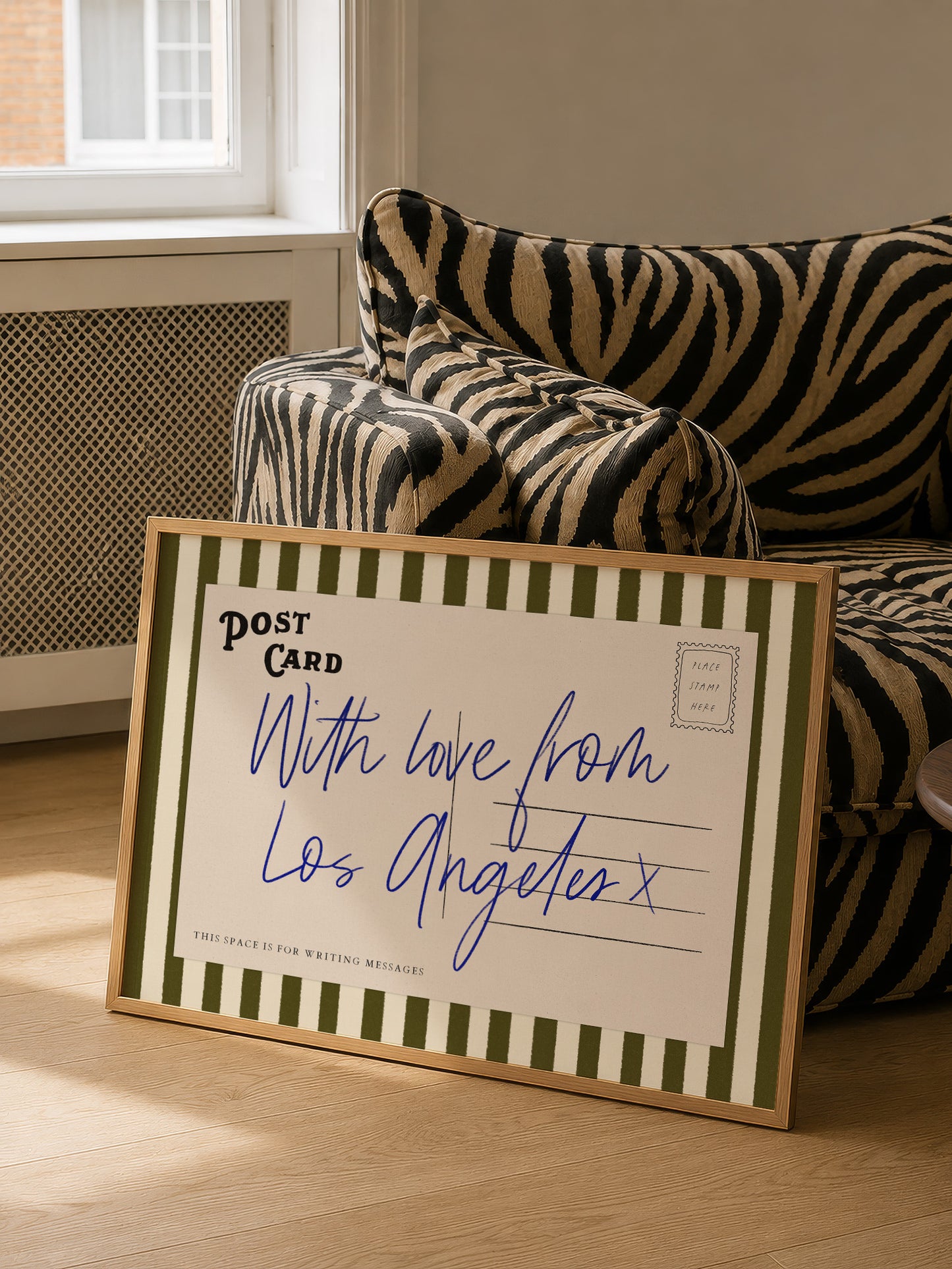A framed postcard with a handwritten message is placed on a couch in a room with a window.