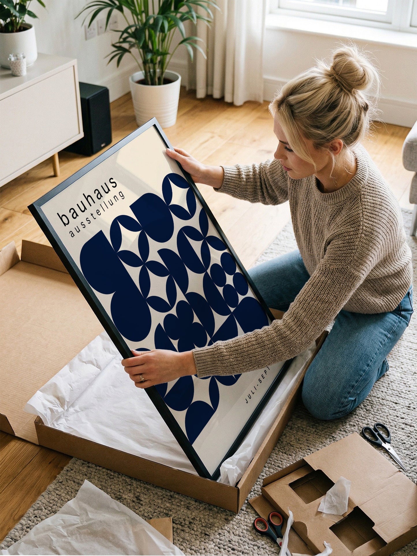 A woman is kneeling on the floor and holding a large framed print with a blue and white geometric design.