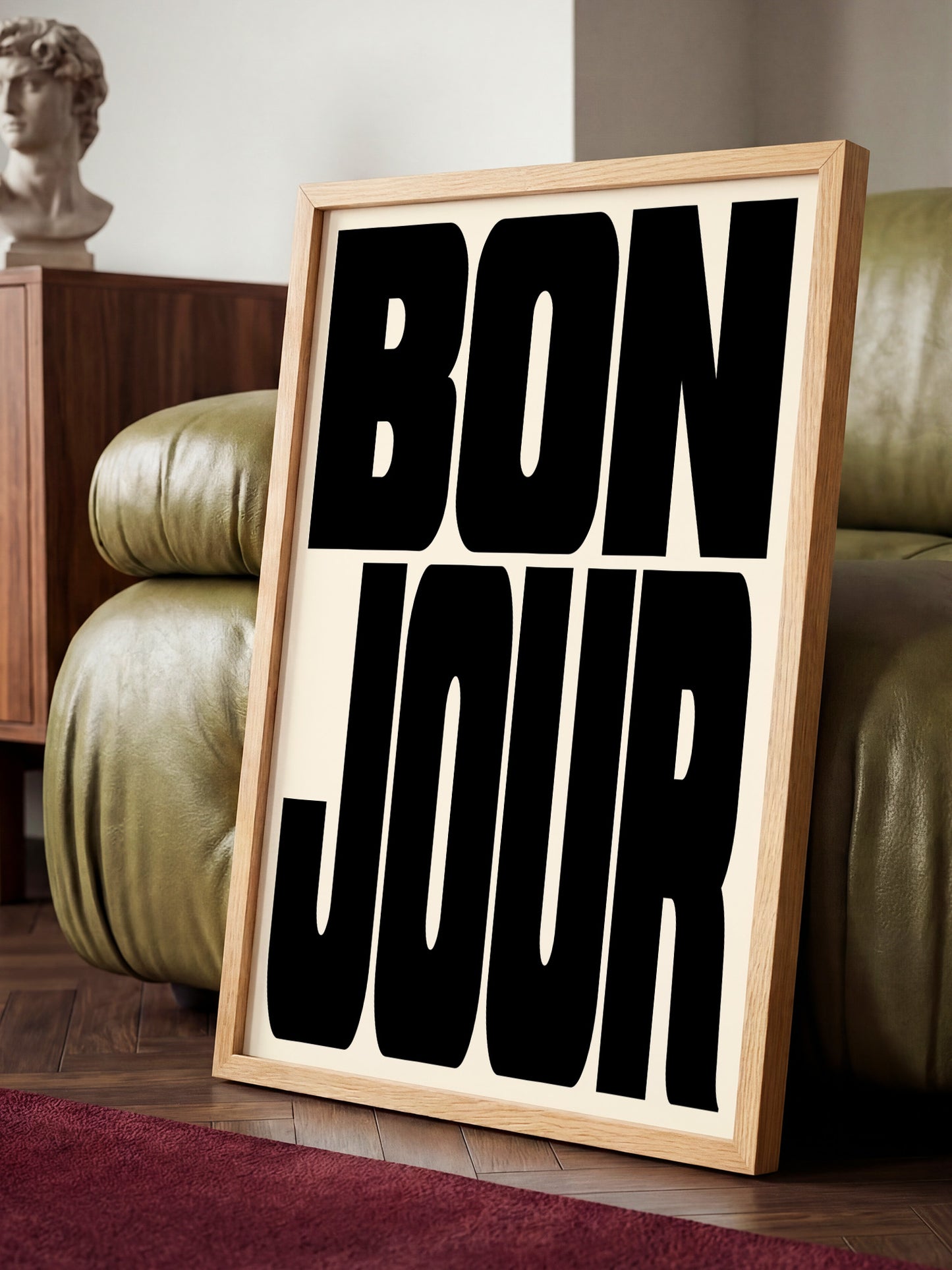 A framed poster with the text " bon jour" in bold black letters is displayed on a wooden floor in a room with a green leather couch.