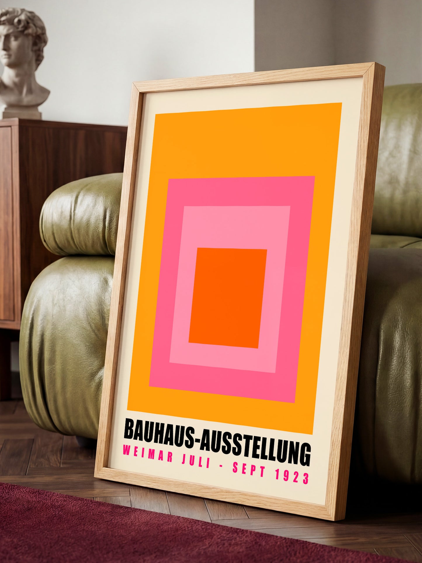 A framed poster with a bold geometric design in orange, pink, and white, sitting on a wooden floor in front of a green leather couch.