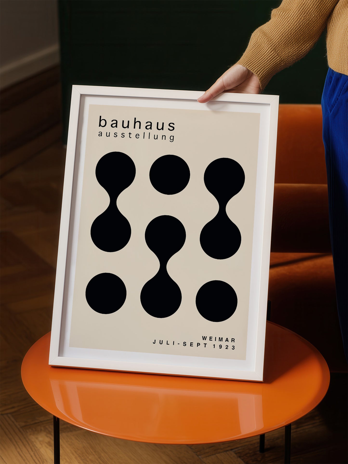 A person is holding a framed poster with a minimalist design featuring black circles on a beige background.