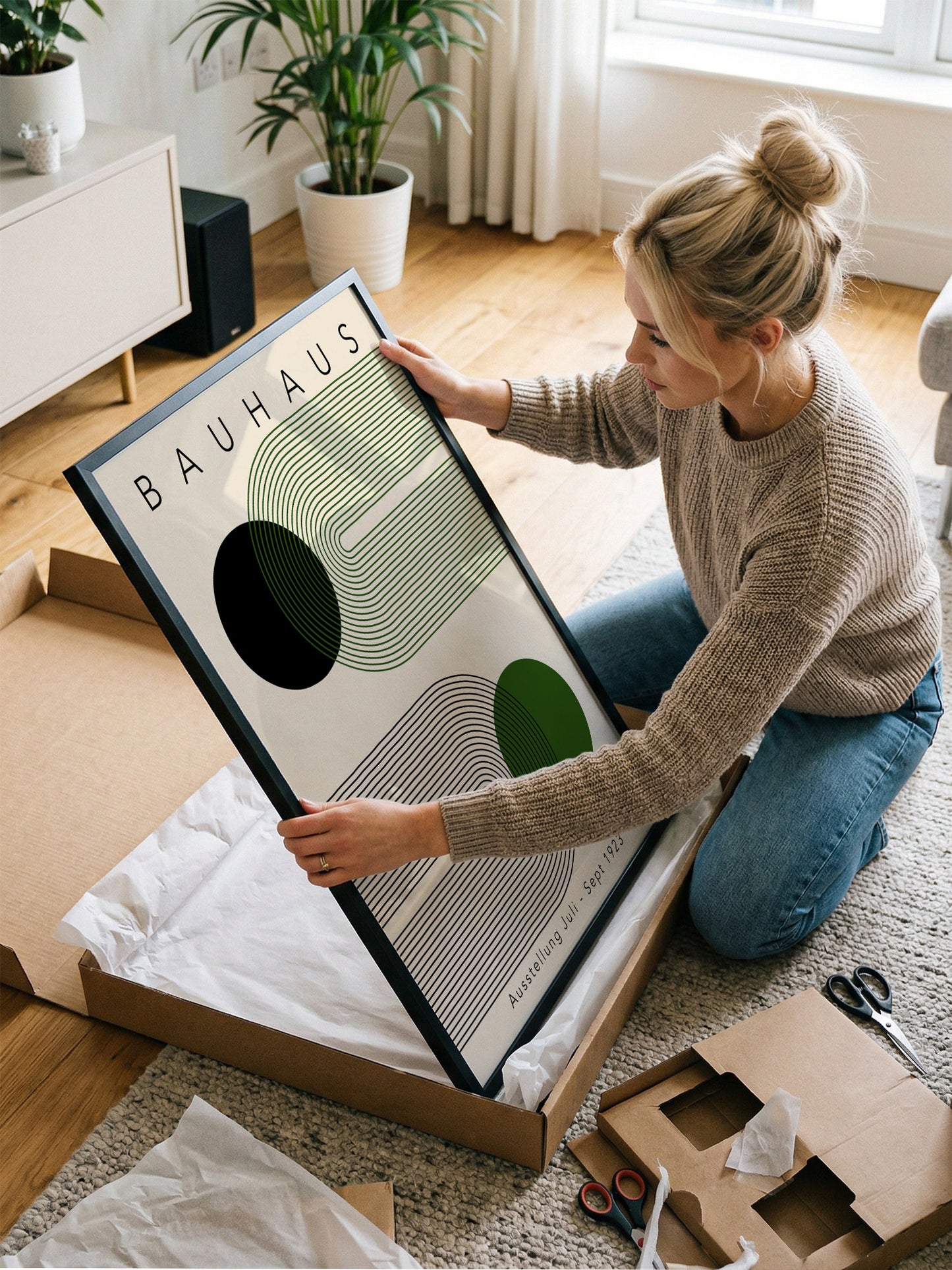 A woman is kneeling on the floor and holding a framed print of the Bauhaus art style.