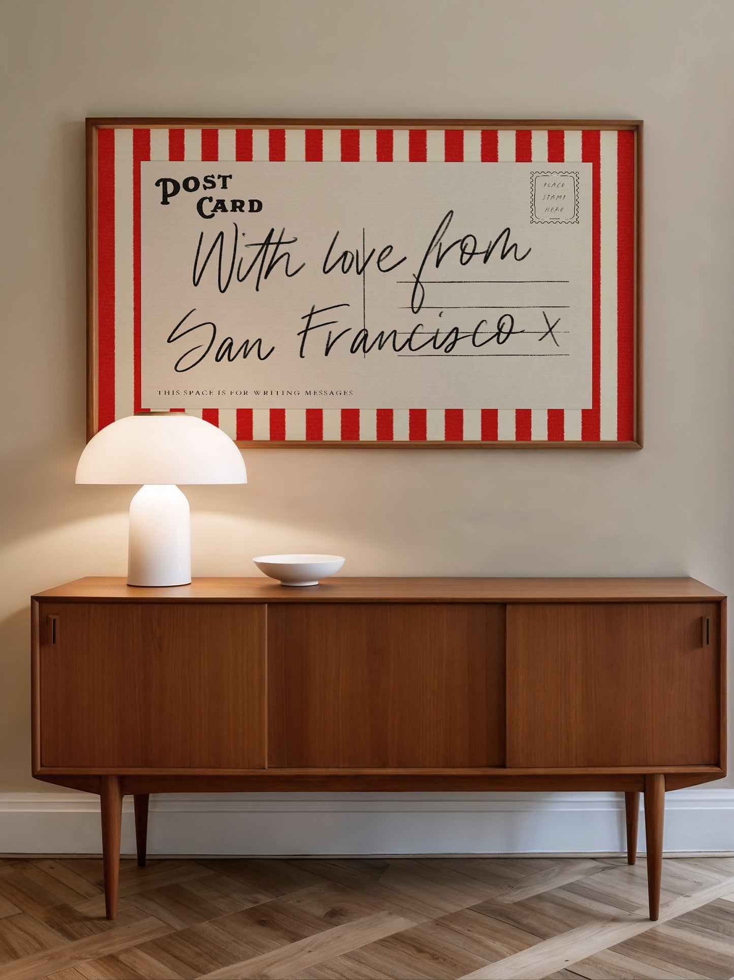 a wooden cabinet with a white lamp on top, and a framed postcard hanging on the wall above it.