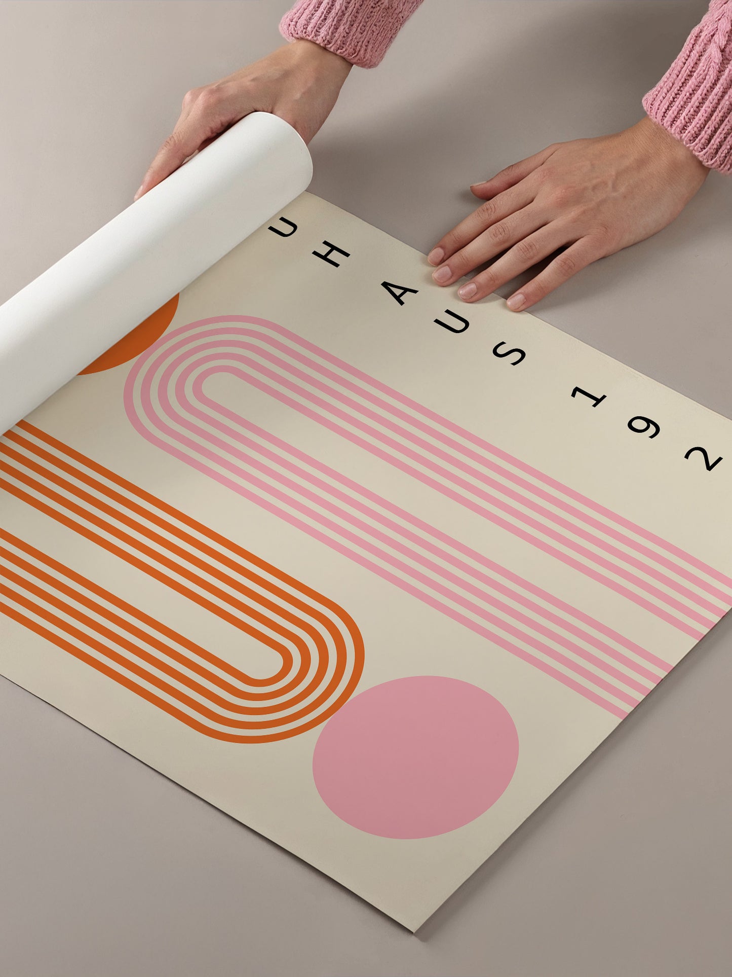 A person's hands are holding a roll of paper, which appears to be a poster or artwork with a geometric design in shades of pink and orange.