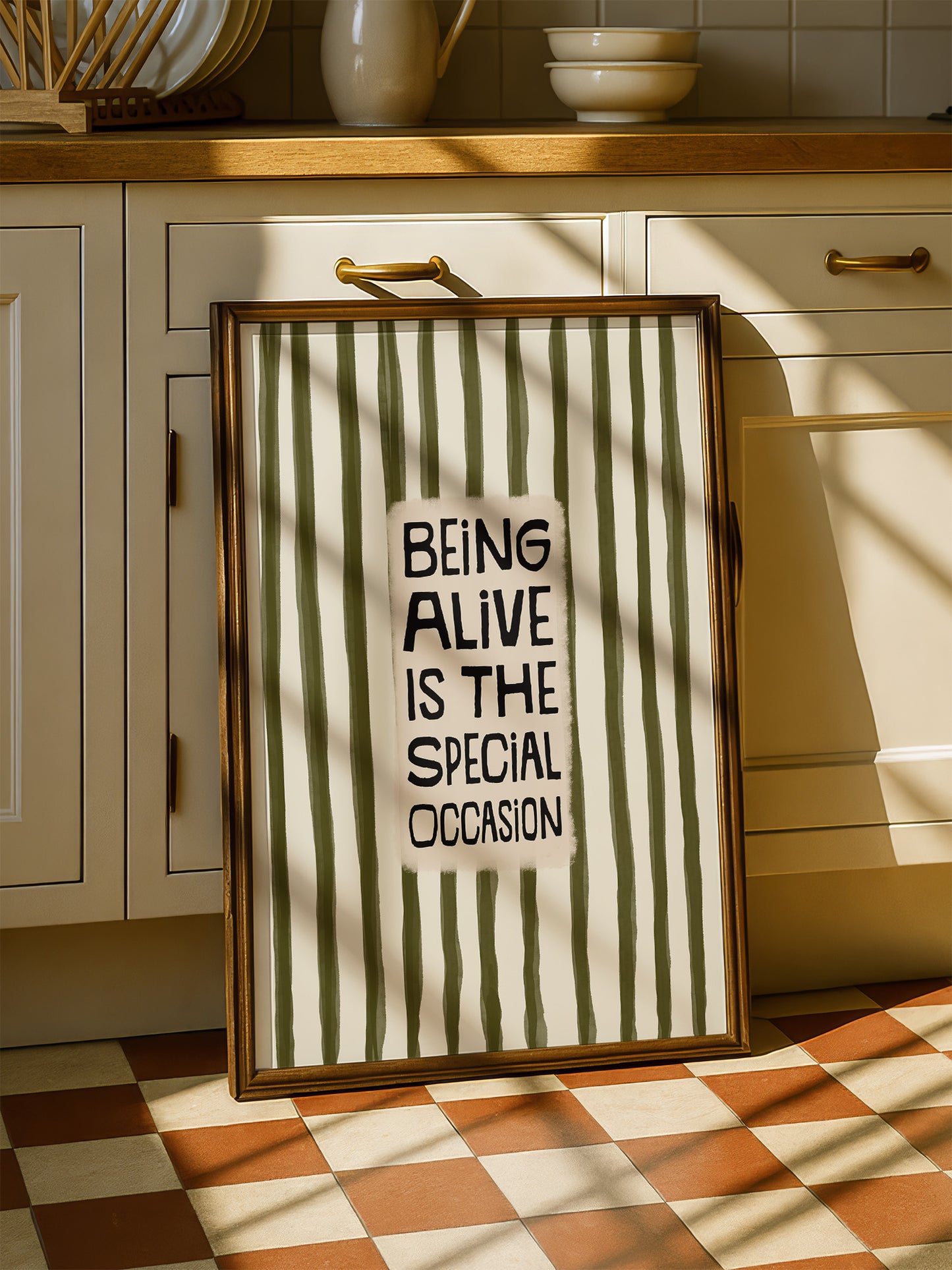 A framed poster with the text "BEING ALIVE IS THE SPECIAL OCCASION" is displayed on a checkered floor in a kitchen setting.