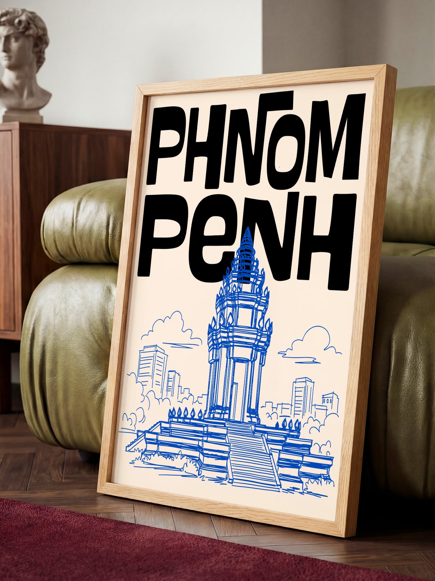 A framed poster with the text "PHNOM PENH" and an illustration of a temple or monument, placed on a wooden floor in front of a green leather couch.