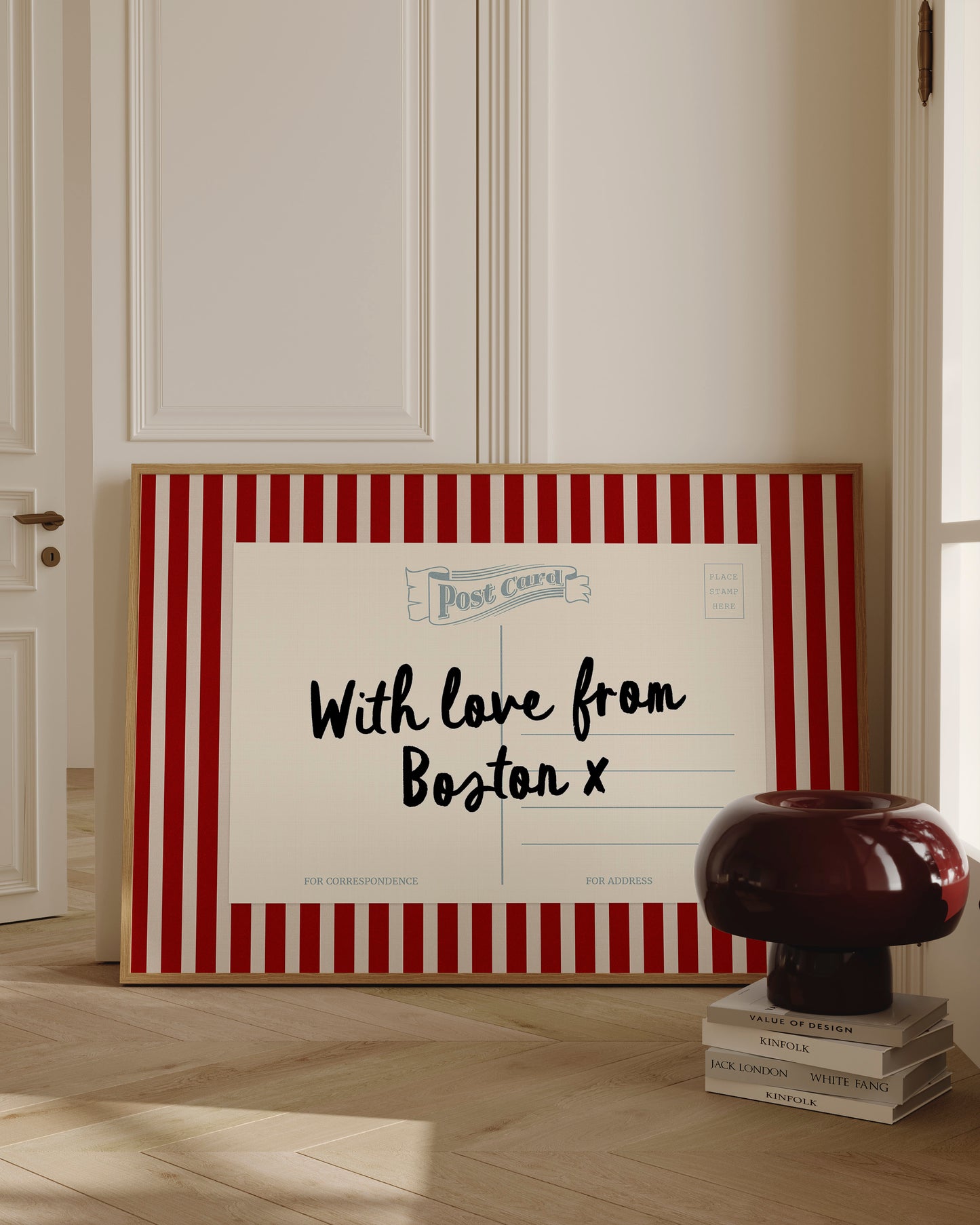A framed postcard with a red and white striped border, displaying a message that reads "With love from Boston X" and "FOR CORRESPONDENCE". The postcard is placed on a wooden floor, with a stack of books and a lamp nearby.