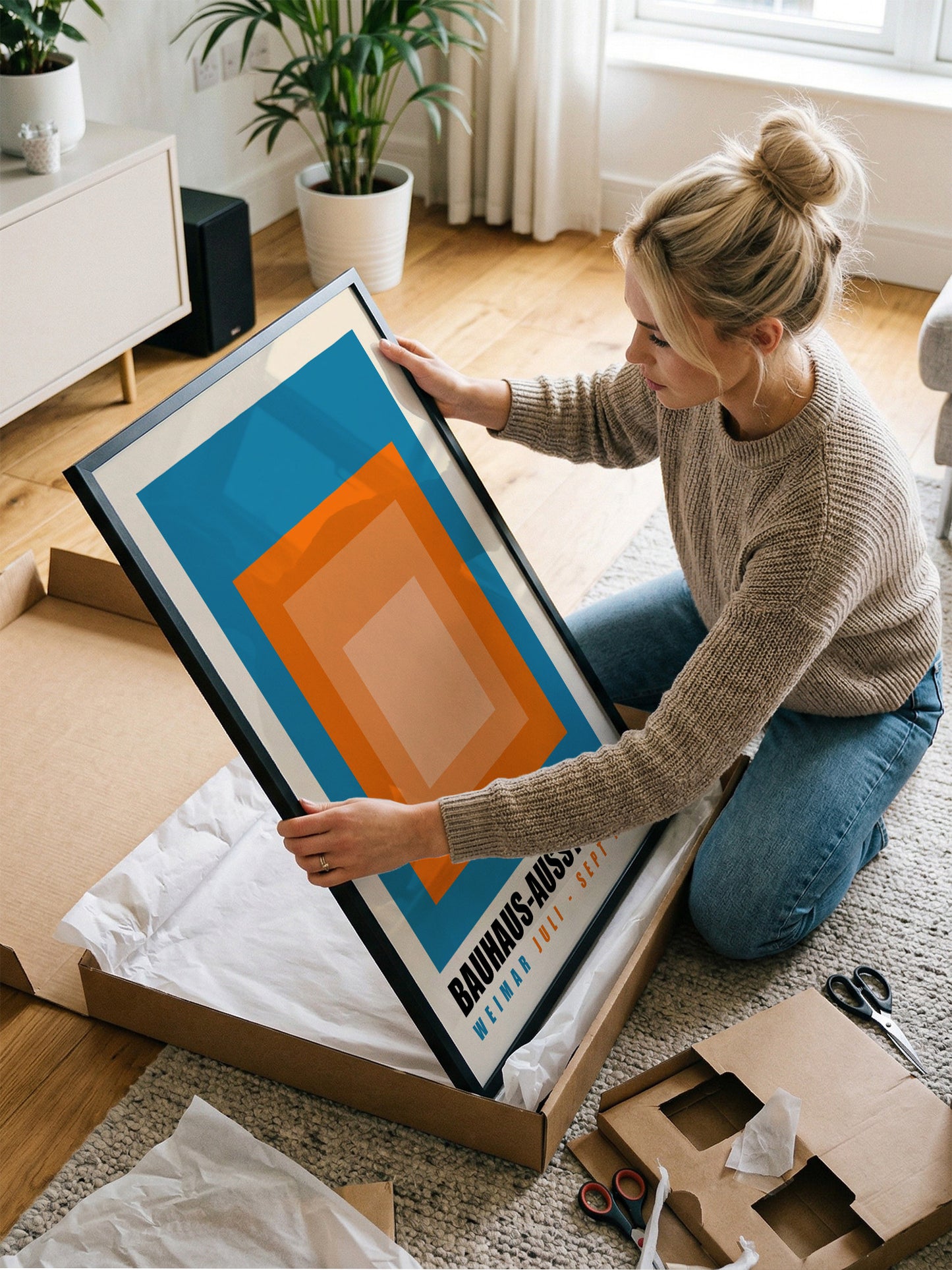 A woman is kneeling on the floor and carefully handling a large framed print, which appears to be a piece of artwork.