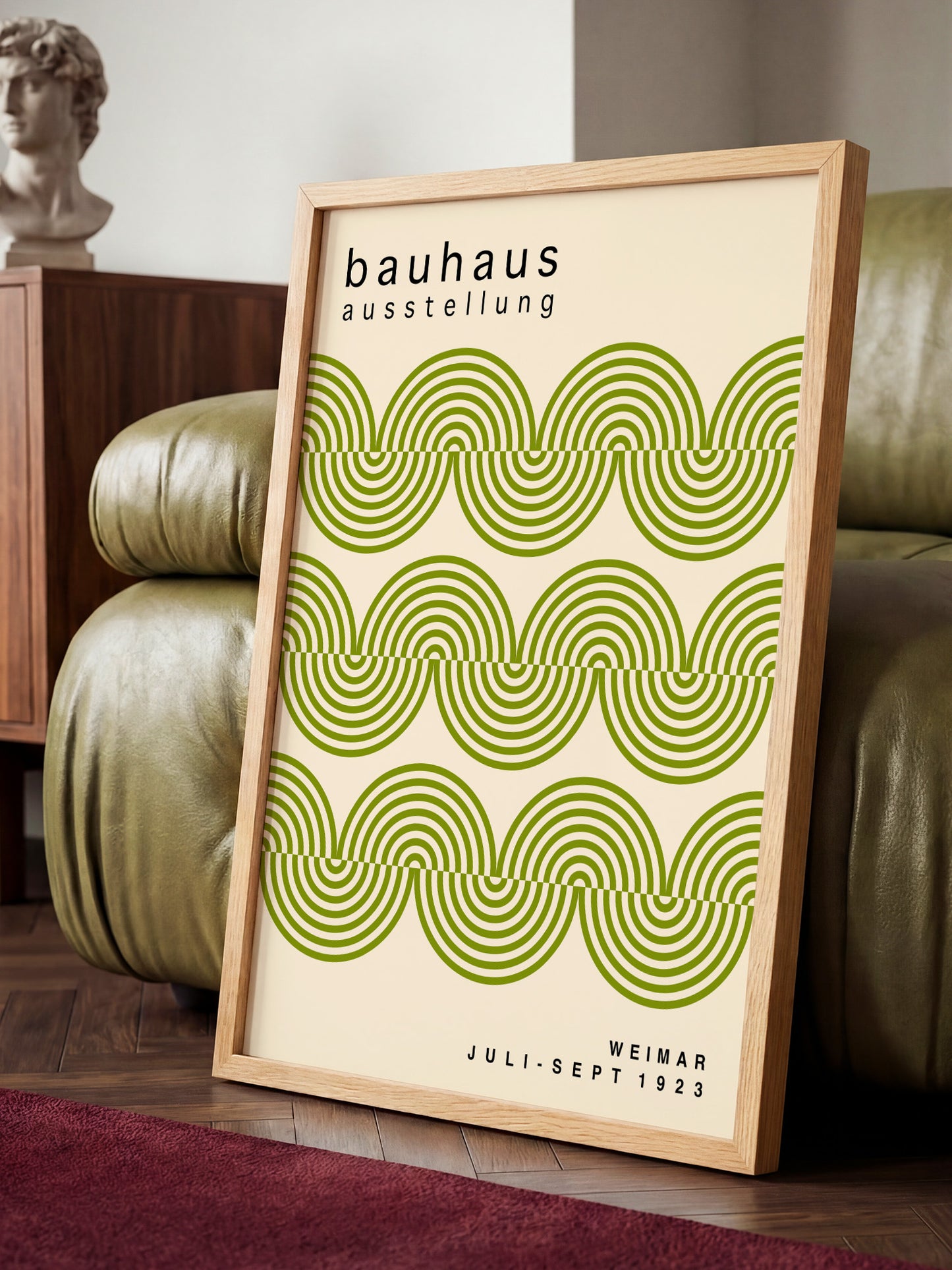 a framed poster of the Bauhaus art school, with a repeating pattern of green and white swirls. The poster is displayed on a wooden floor, with a green leather couch in the background.