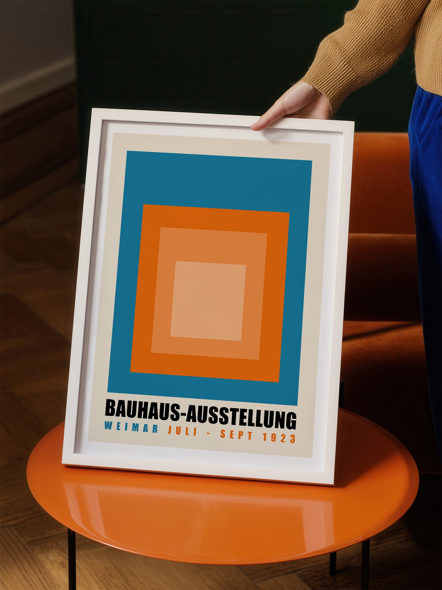 A person is holding up a framed poster with a bold geometric design in blue, orange, and white colors.
