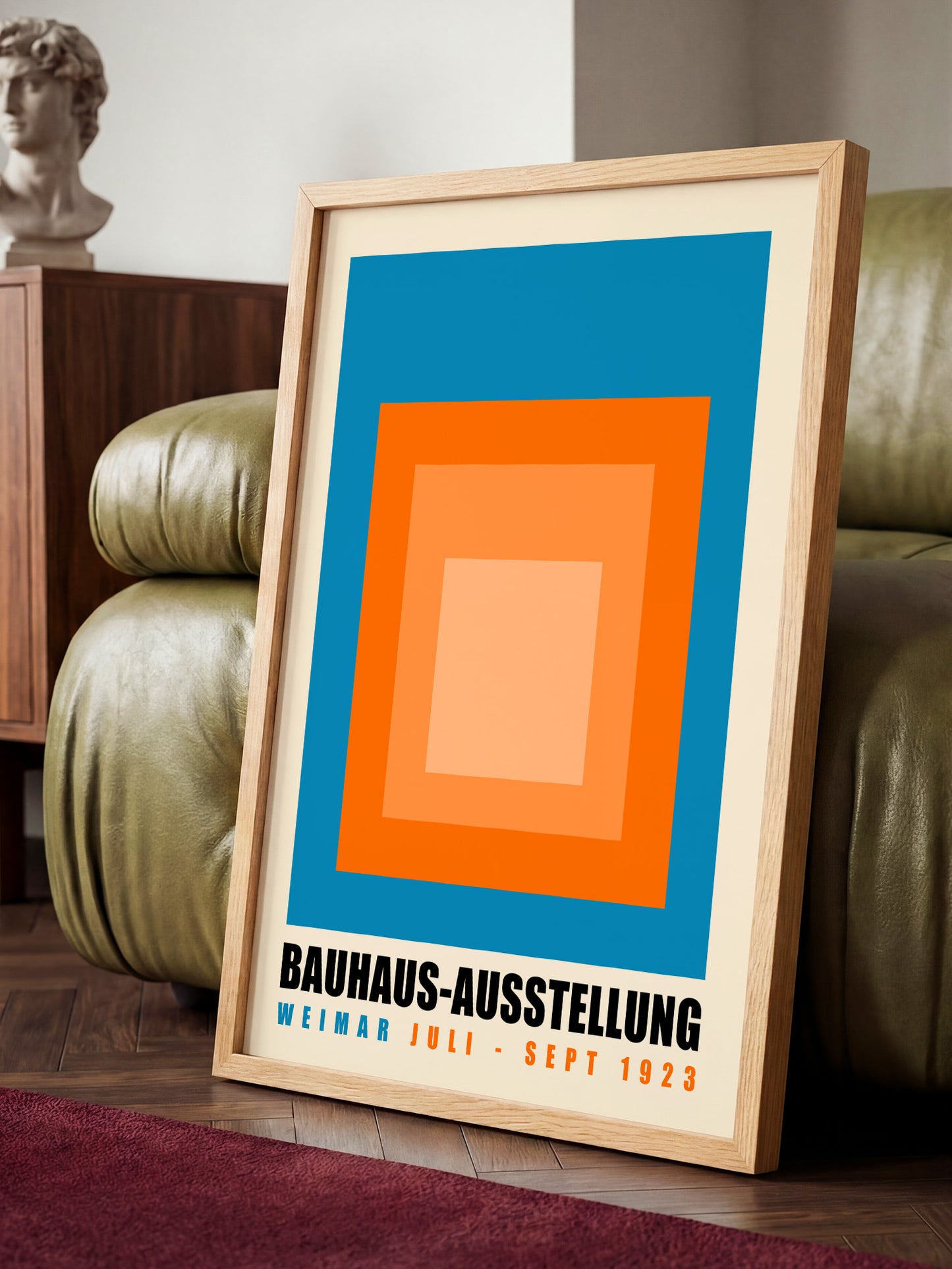 A framed poster with a bold geometric design in blue, orange, and white colors, sitting on a wooden floor in front of a green leather couch.