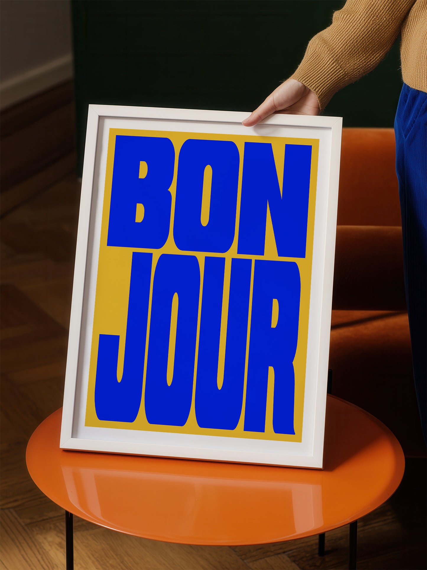 A person is holding up a framed poster with the text "BON JOUR" in large blue letters on a yellow background.