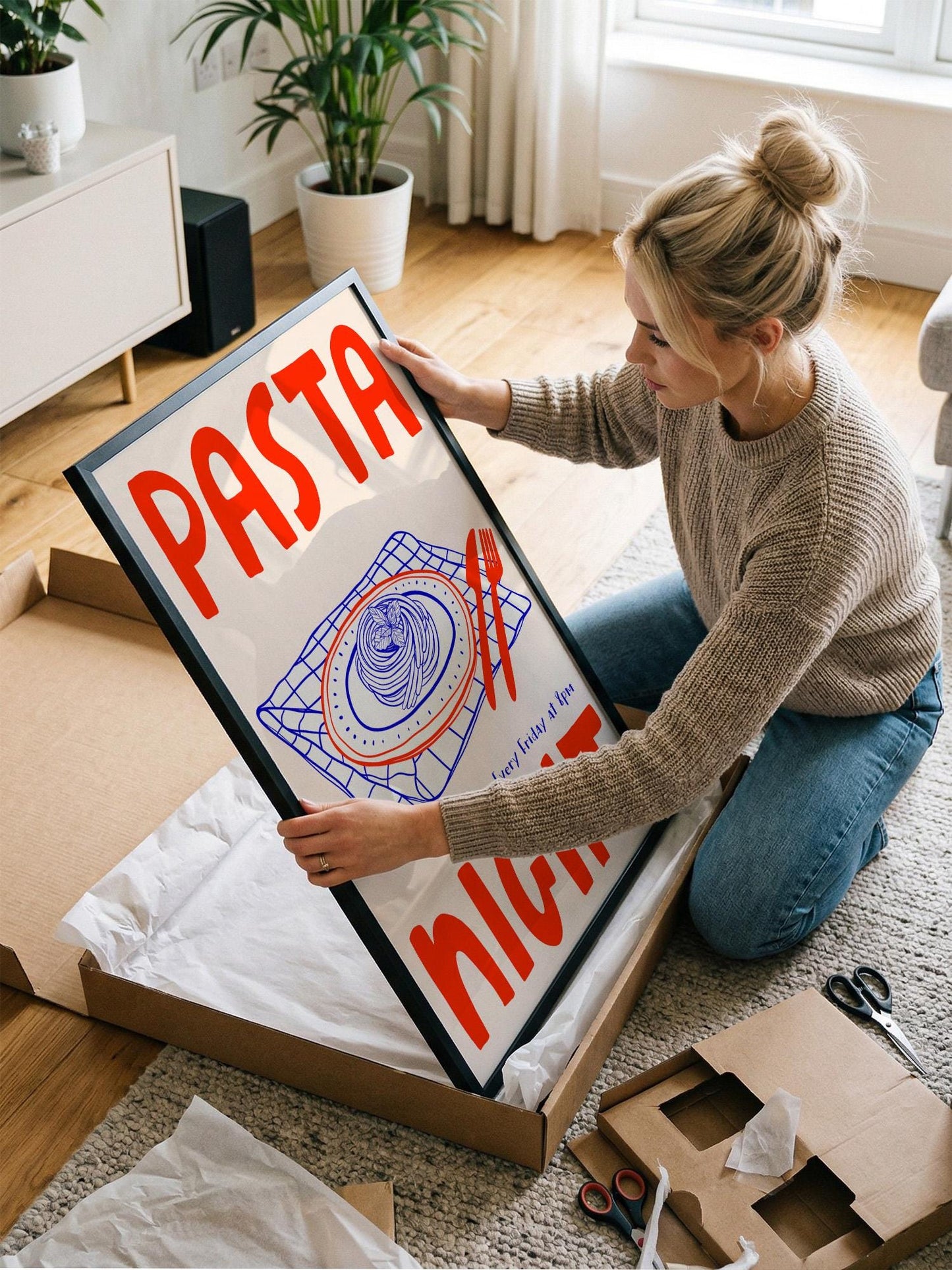 A woman is kneeling on the floor and holding a framed poster with the word "PASTA" on it.