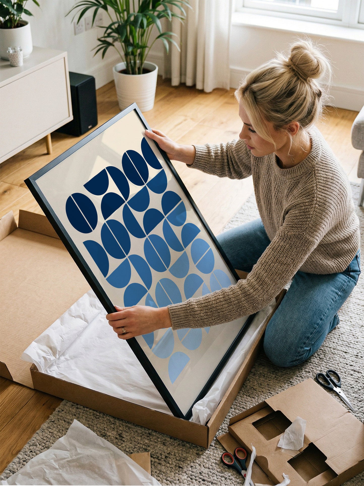A woman is kneeling on the floor, carefully opening a large cardboard box containing a framed artwork with a geometric pattern in shades of blue and white.