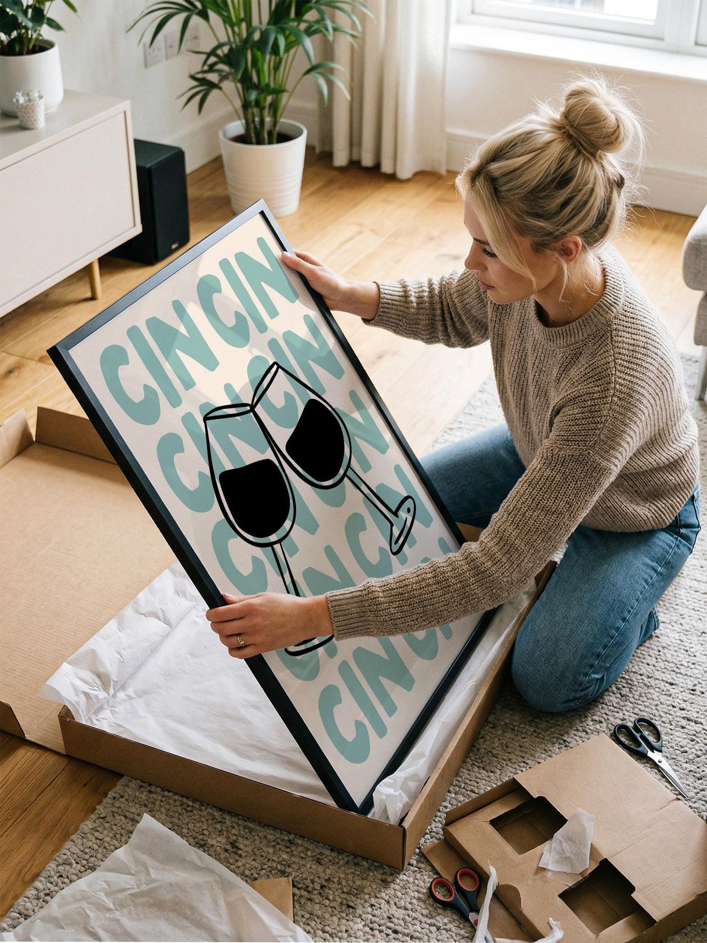 A woman is kneeling on the floor and holding a framed poster with a wine glass design.