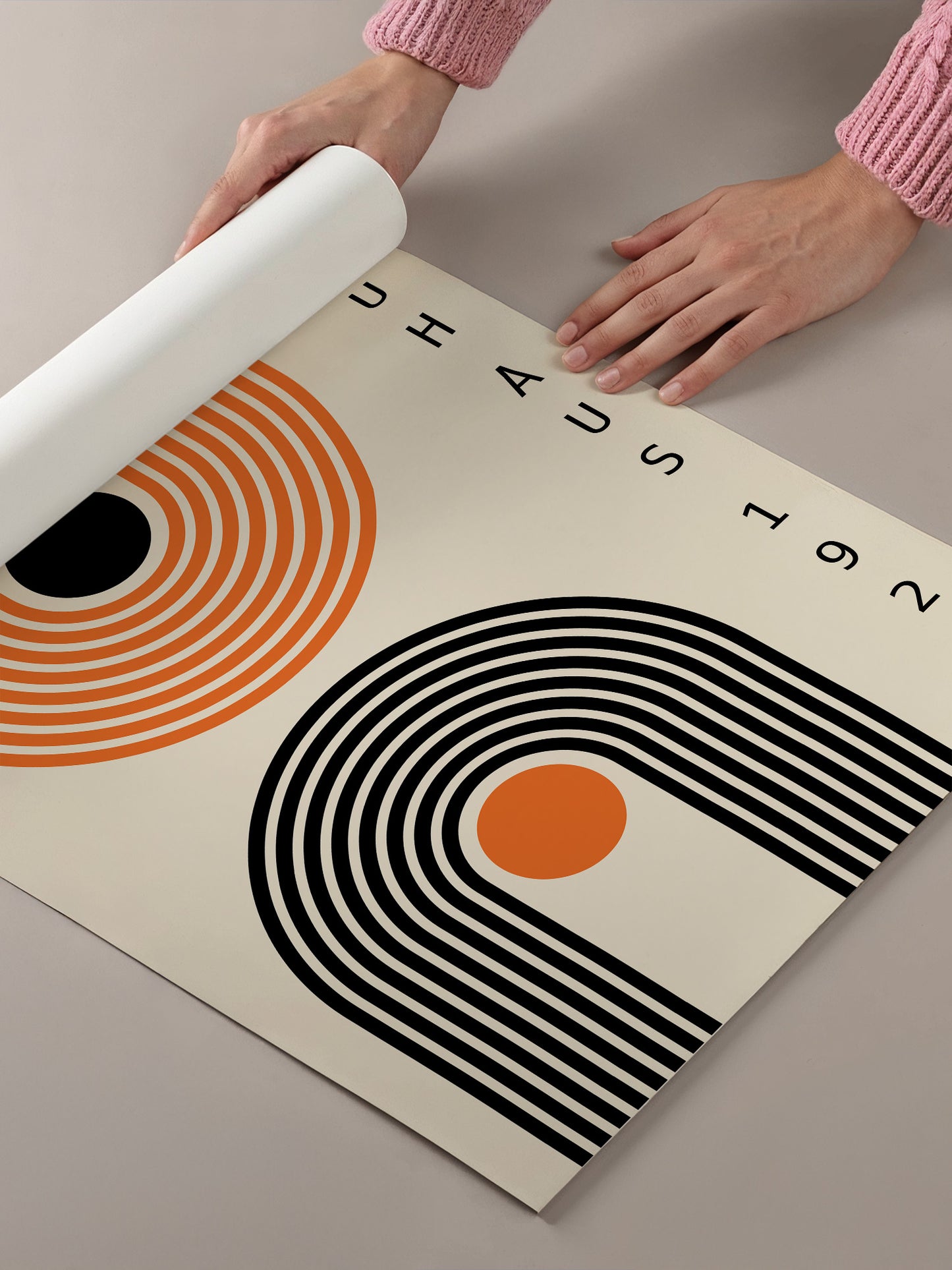 A person's hands are holding a roll of paper, preparing to print a poster with a geometric design featuring black and orange lines and a central orange circle.