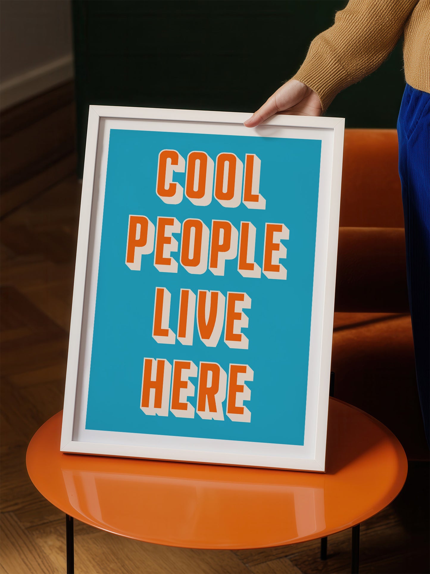 A person is holding up a framed poster with the text "COOL PEOPLE LIVE HERE" in a blue background.
