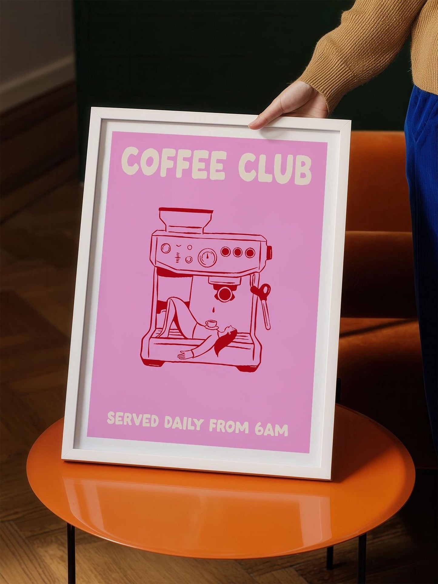 A person is holding a framed poster with a pink background and a cartoon illustration of a coffee machine. The poster has the text "COFFEE CLUB" and "SERVED DAILY FROM 6AM" printed on it.
