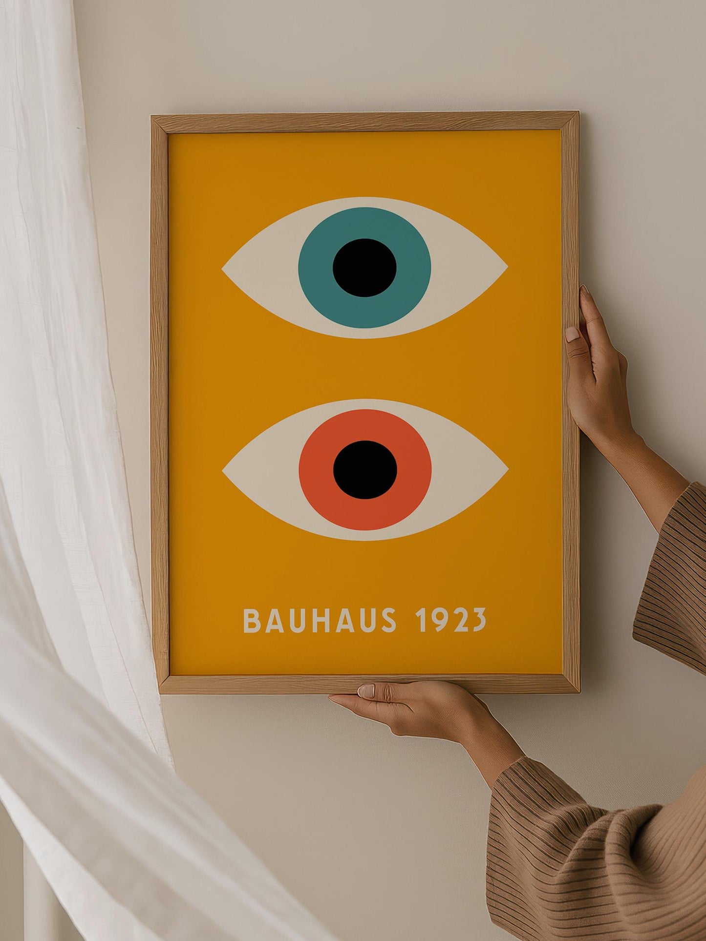 A person is holding up a framed poster with a bold yellow background and two large eye-like symbols, one blue and one red, against a white wall.
