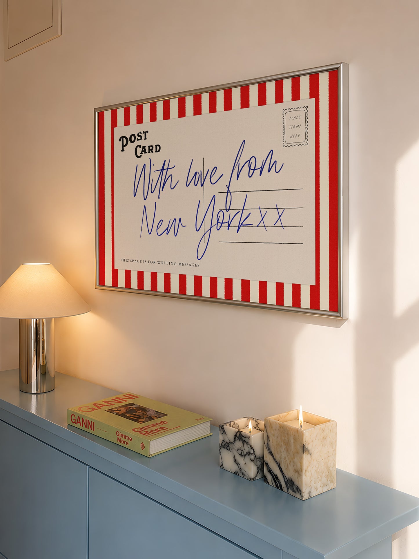 a framed postcard with a handwritten message, a lamp, a book, and a marble candle holder on a blue dresser.