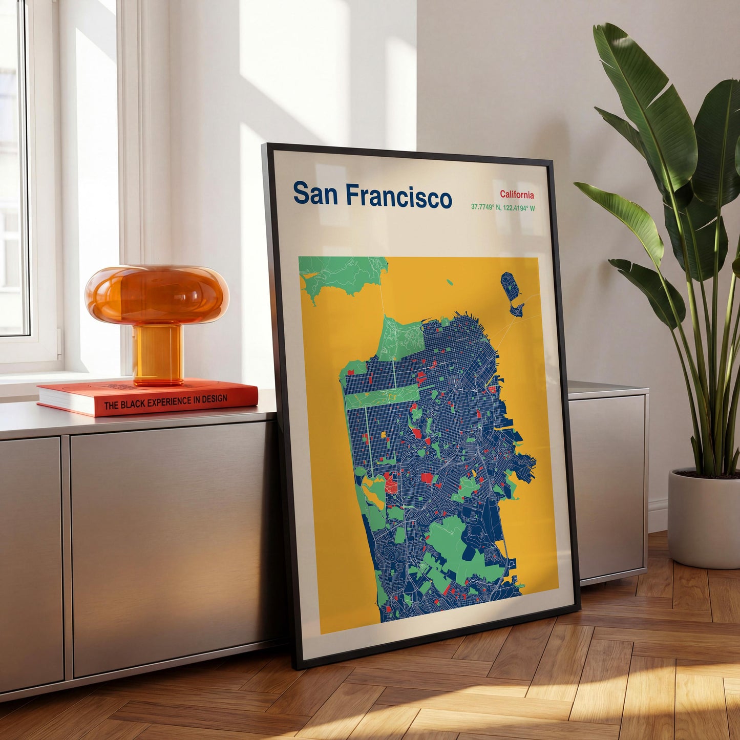 a framed map of San Francisco, California, with a vibrant color scheme of yellow, blue, and green. The map displays the city's streets and neighborhoods, providing a visual representation of the area. The frame is placed on a wooden floor, and there is a potted plant nearby.