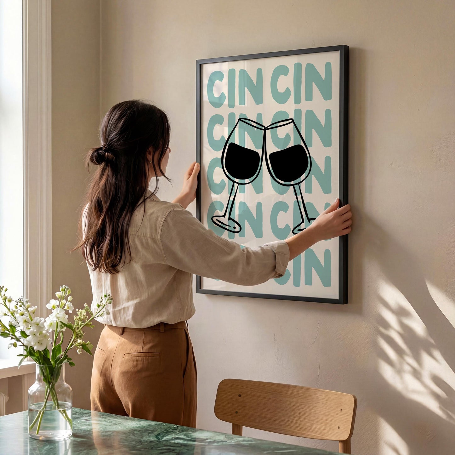 A woman is hanging a framed poster on a wall, which features a graphic of two wine glasses clinking together.