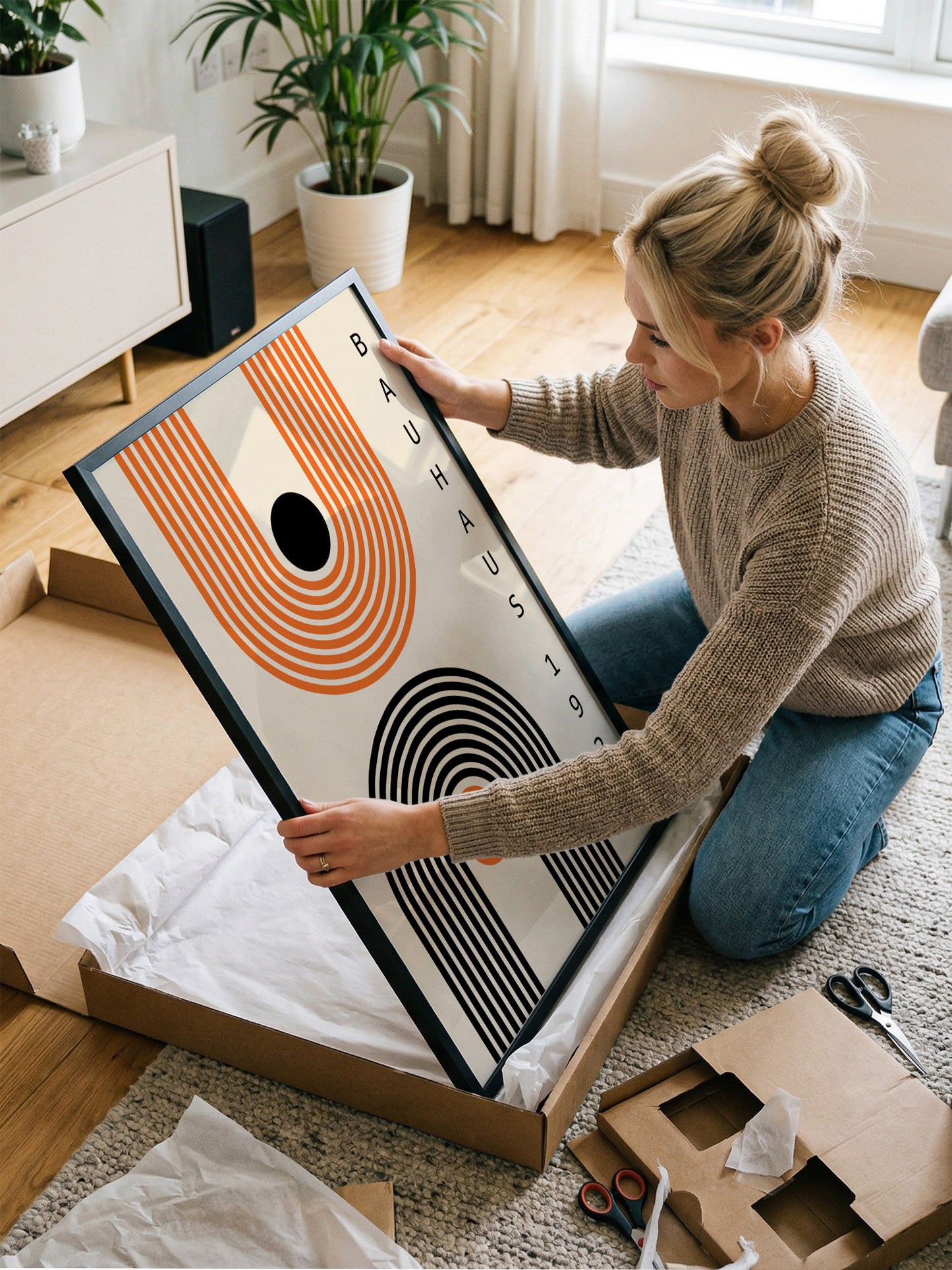 A woman is kneeling on the floor and carefully opening a large picture frame, which is a Bauhaus-style abstract artwork.