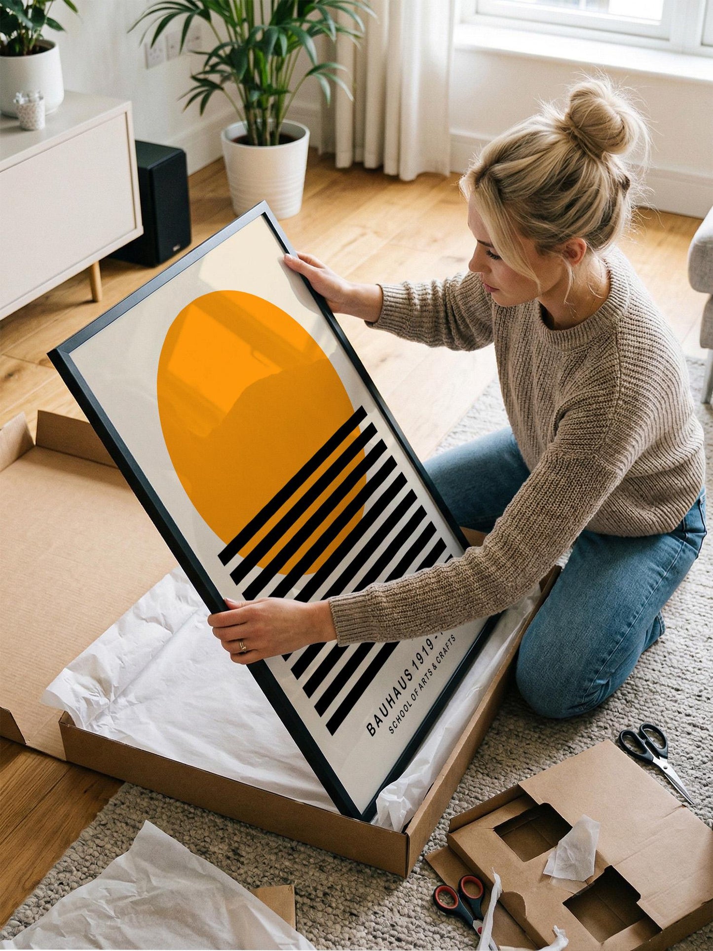 A woman is kneeling on the floor and carefully opening a large cardboard box, which contains a framed artwork.