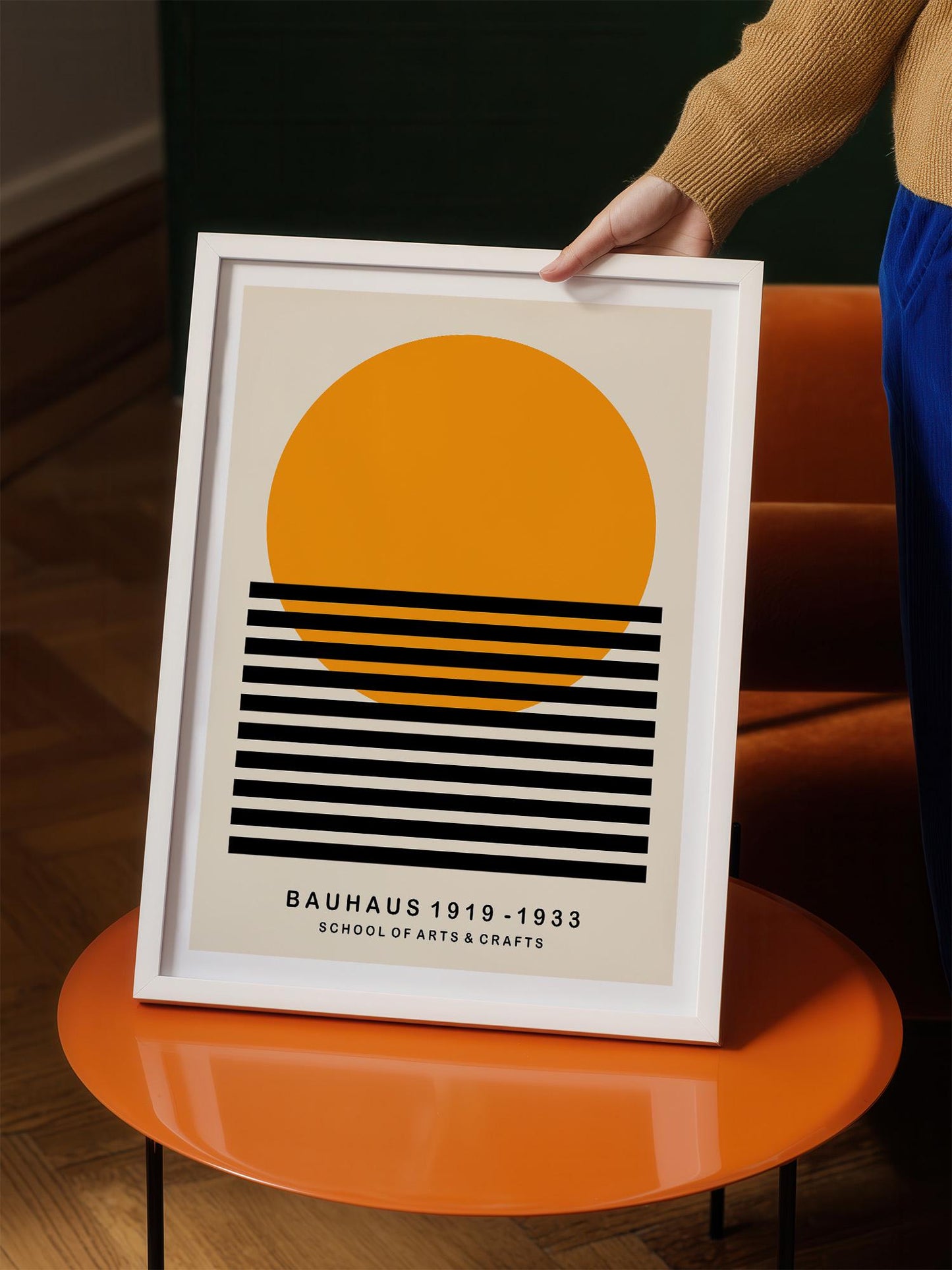 A person is holding up a framed poster with a minimalist design featuring a large orange circle and black horizontal lines.