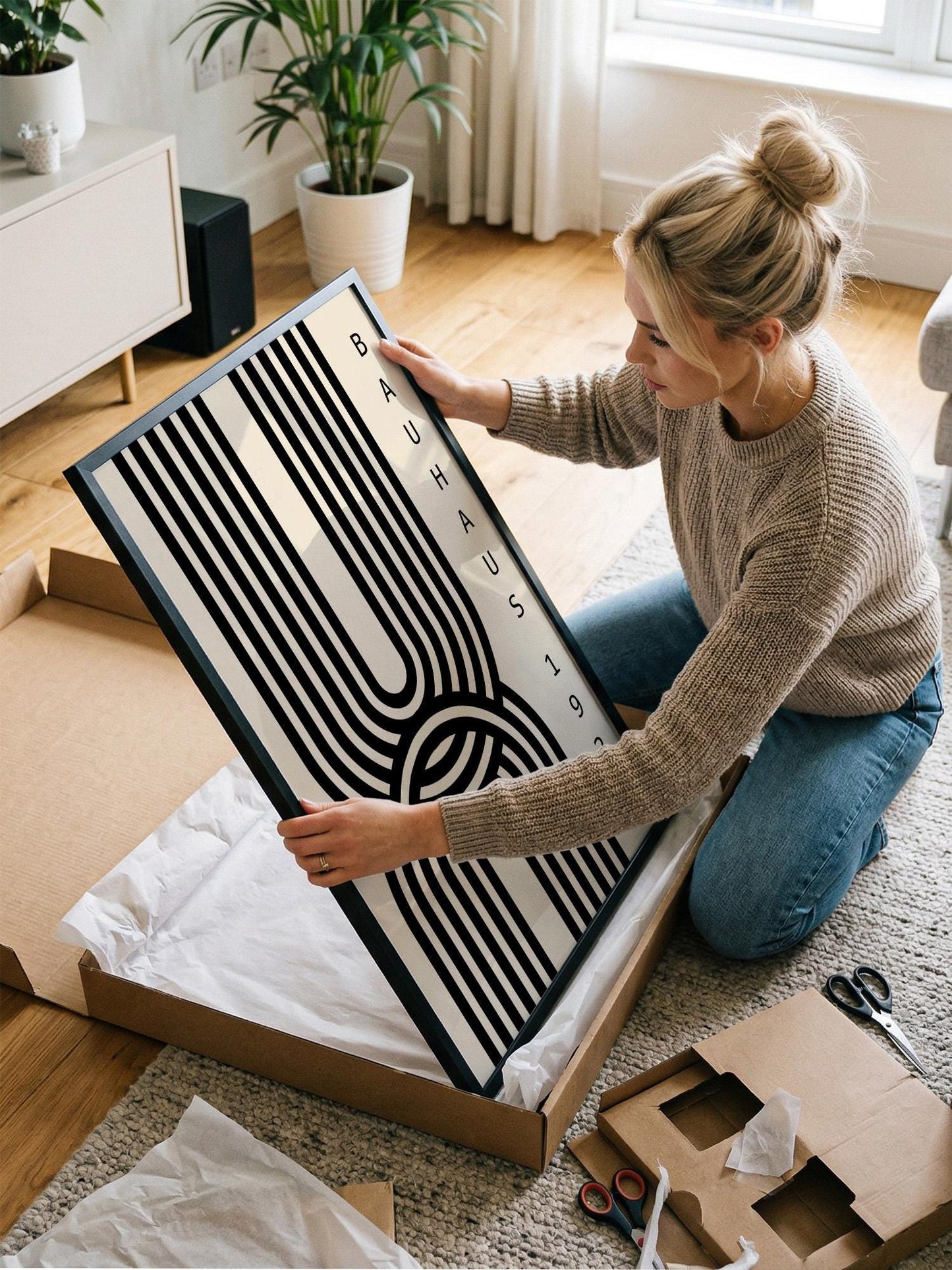 A woman is kneeling on the floor and carefully opening a large cardboard box, which contains a framed artwork.