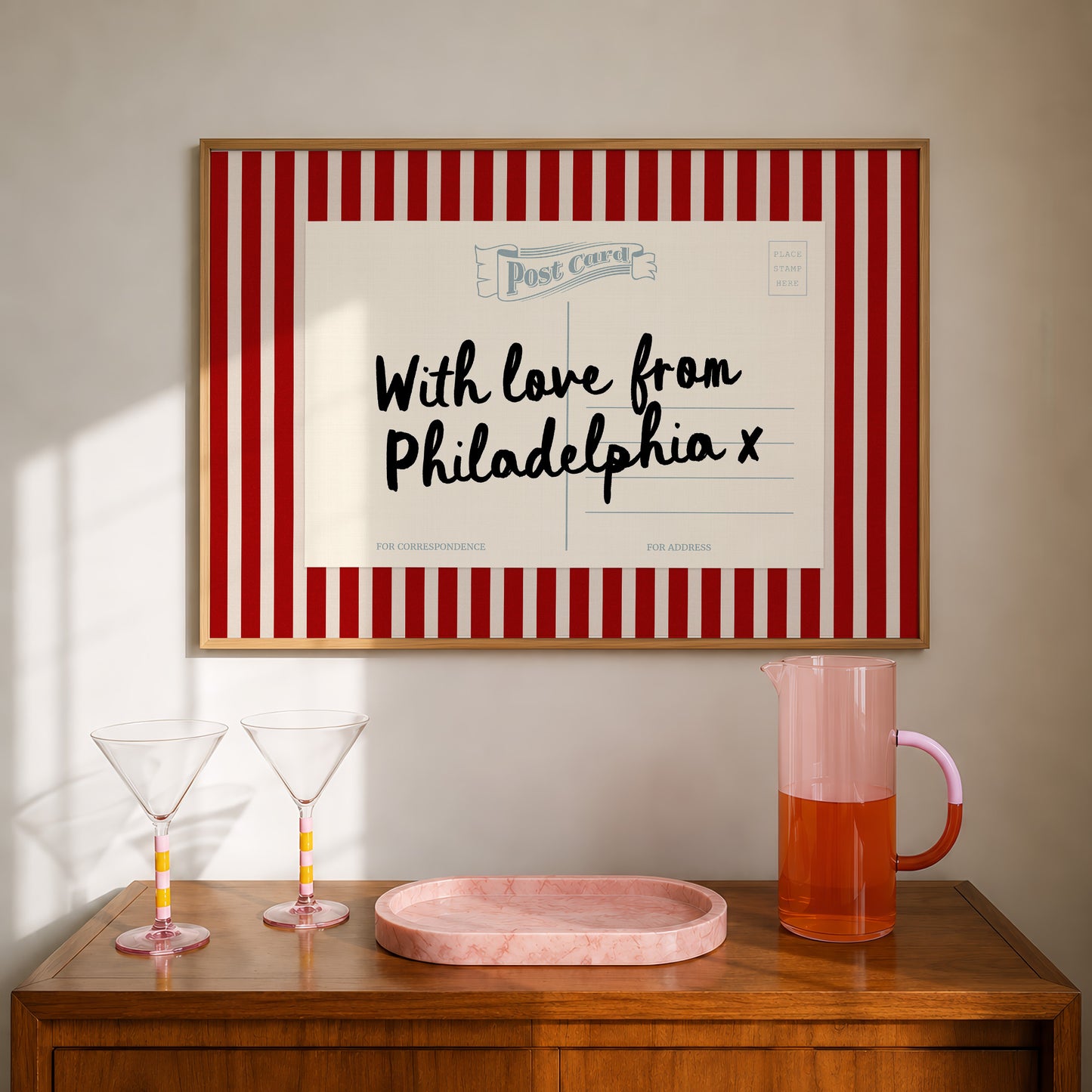 a wooden table with two martini glasses, a pink pitcher, and a framed postcard with a message.