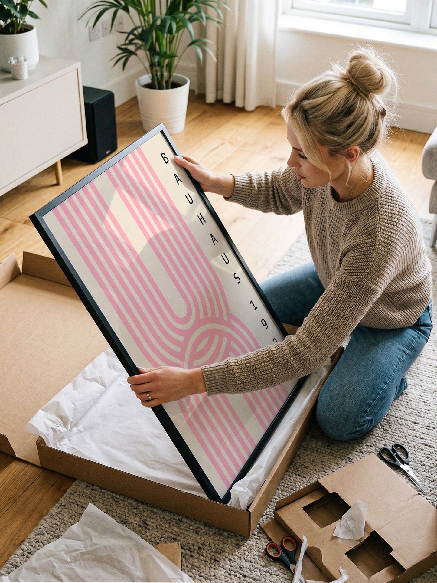 A woman is kneeling on the floor and carefully opening a large cardboard box, which contains a framed poster.