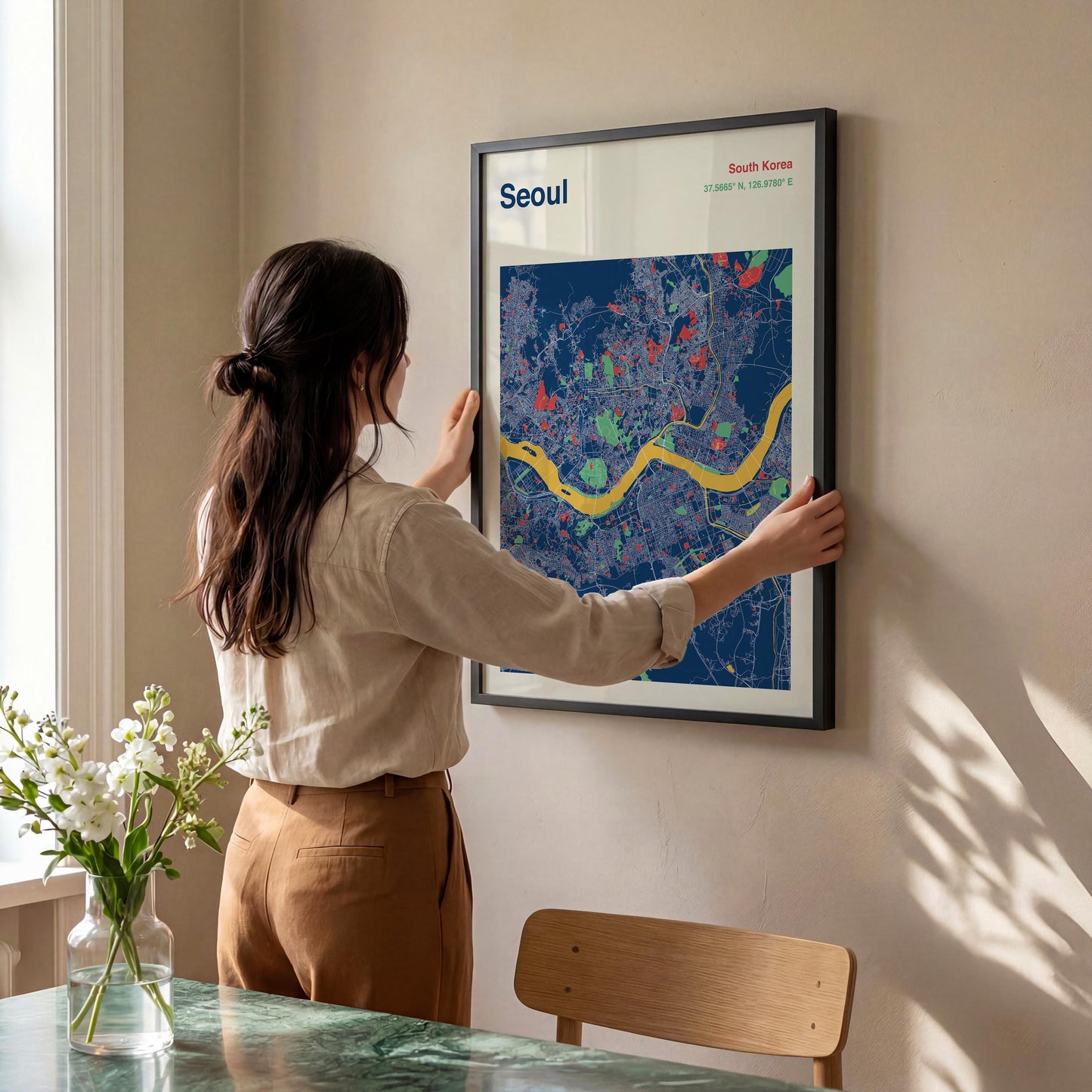 A woman is hanging a framed poster of a map of Seoul, South Korea, on a wall.