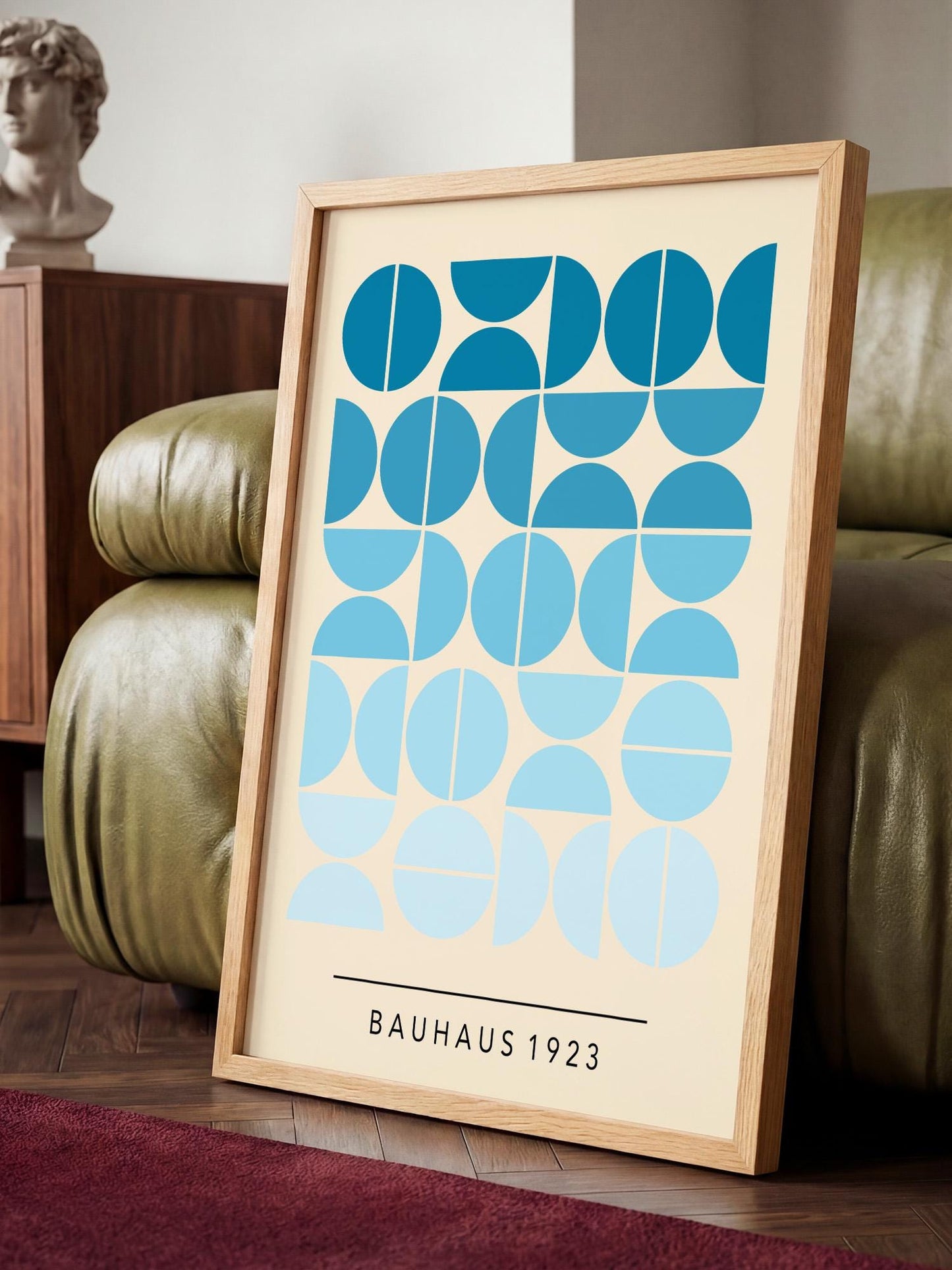 a framed poster with a geometric design of blue circles and lines, placed on a wooden floor in front of a green leather couch.