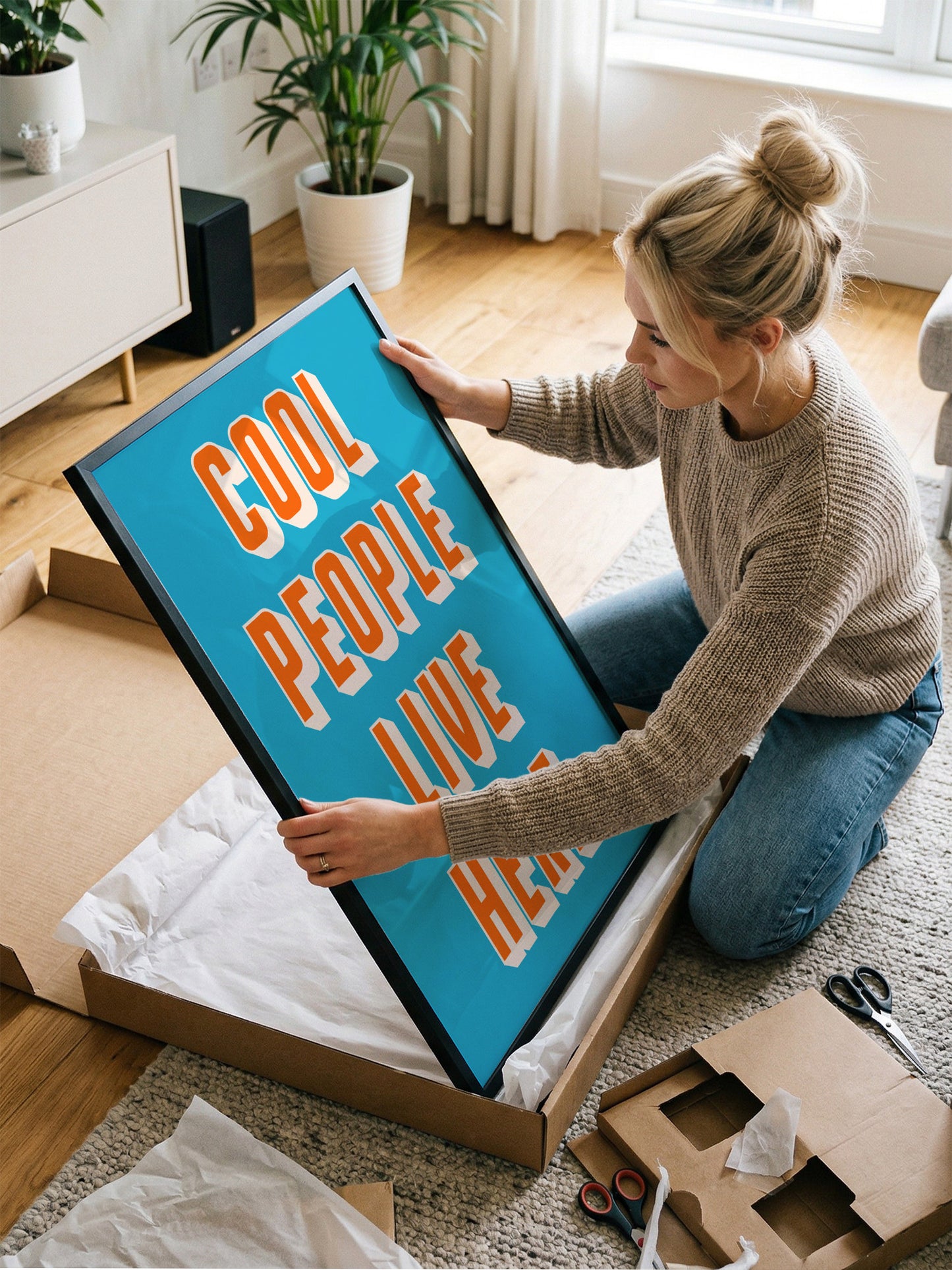 A woman is kneeling on the floor and opening a large cardboard box, which contains a large blue sign with the text "COOL PEOPLE LIVE HERE" written on it.