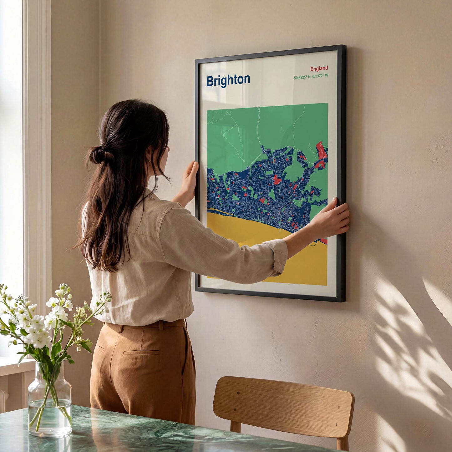 A woman is hanging a framed poster of Brighton on a wall in a room with a dining table and a vase of flowers.