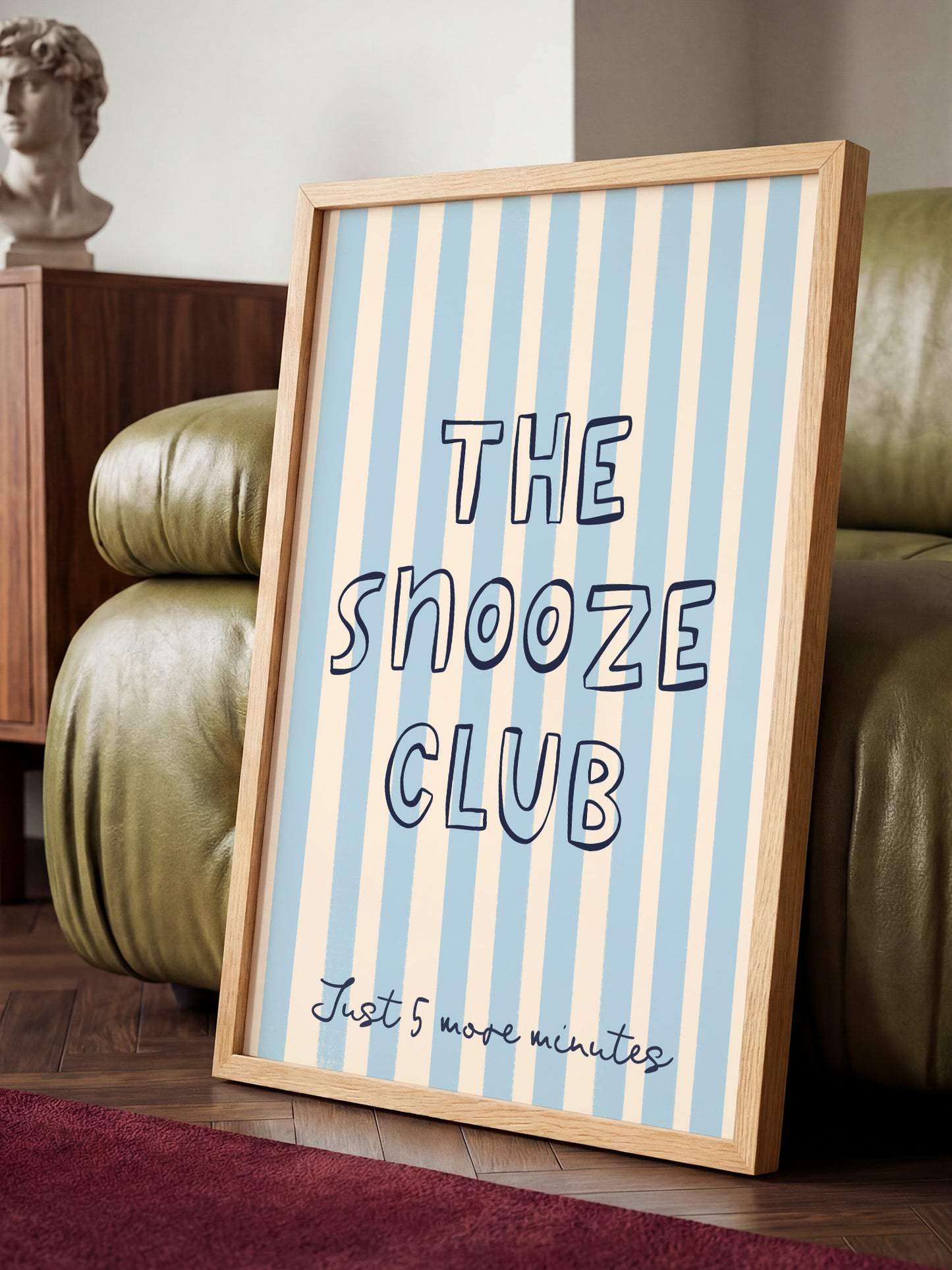 A framed sign with the text "THE SNOOZE CLUB" and "Just 5 more minutes" is displayed on a wooden floor in a room with a green leather couch.