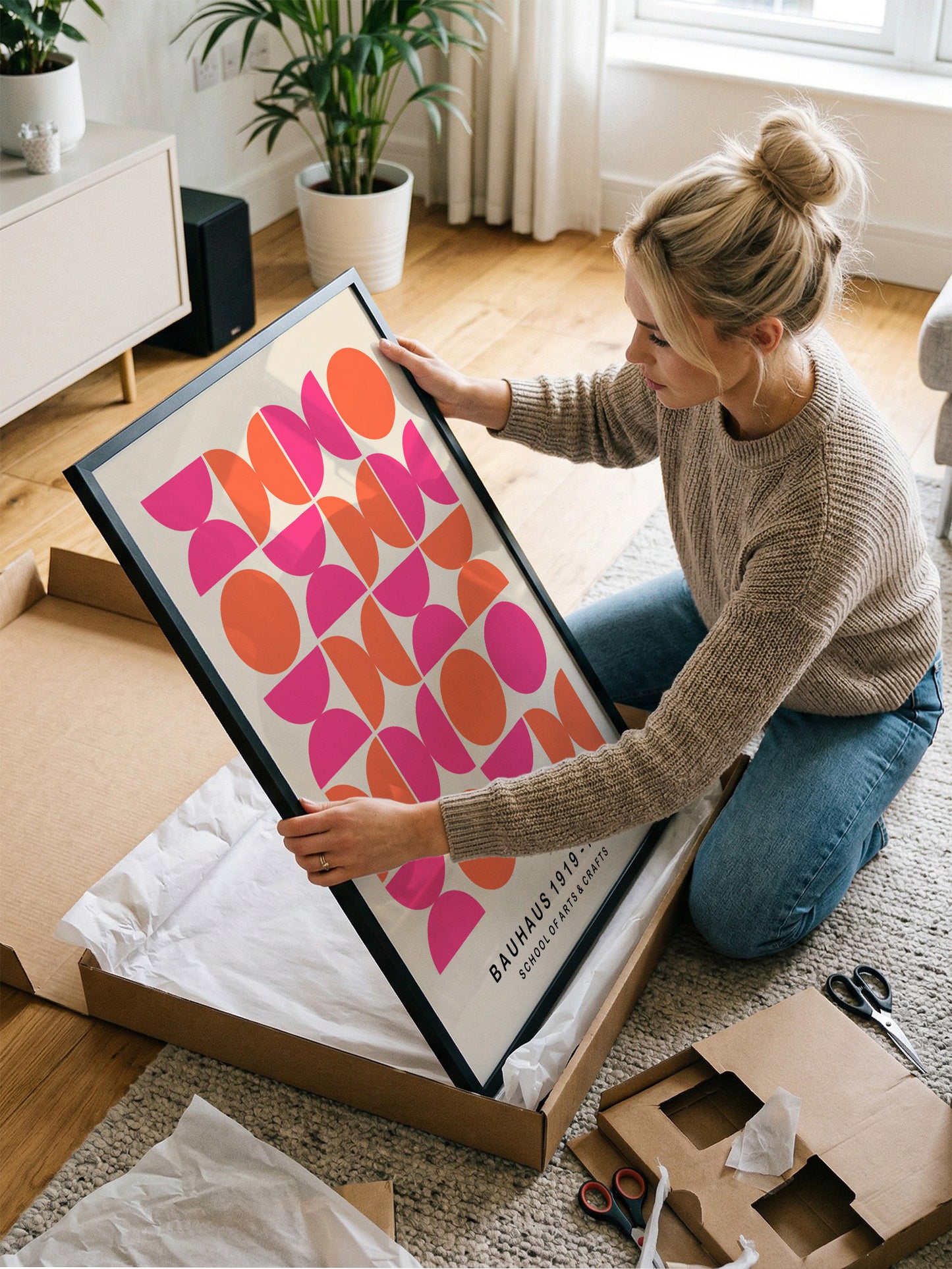 A woman is kneeling on the floor and carefully opening a large cardboard box, which contains a framed poster with a bold geometric design in pink and orange colors.