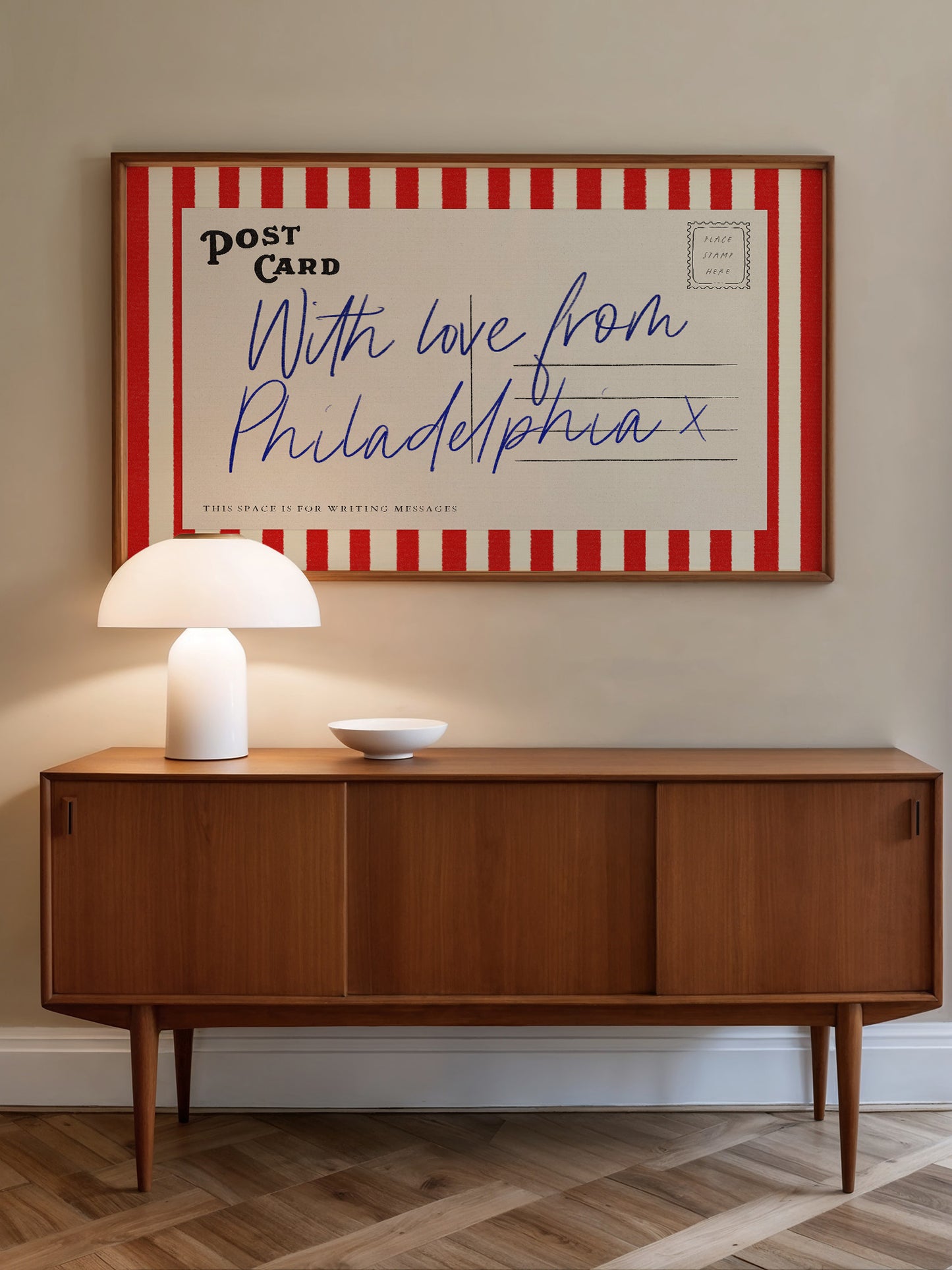 a wooden cabinet with a white lamp on top, and a framed postcard hanging on the wall above it.