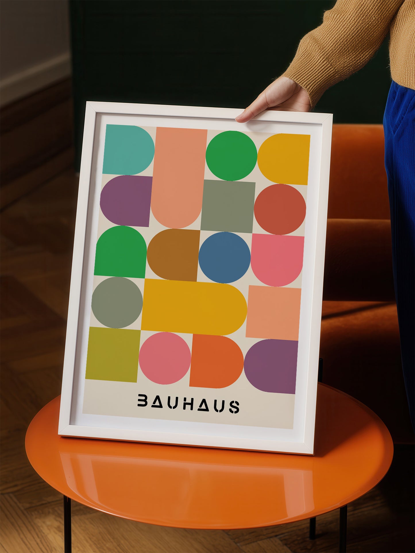 A person is holding up a framed poster with a geometric design featuring various shapes and colors, including circles, triangles, and rectangles.