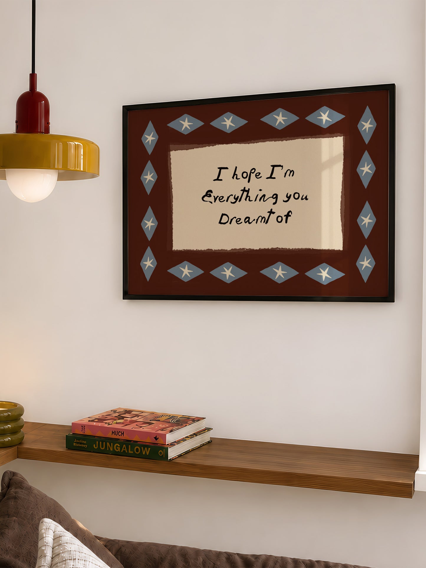 a framed wall art with a message that reads "I hope I'm Everything you Dreamt of". The art is framed with a blue and white diamond pattern and hangs on a white wall above a wooden shelf. There are also some books on the shelf below the art.