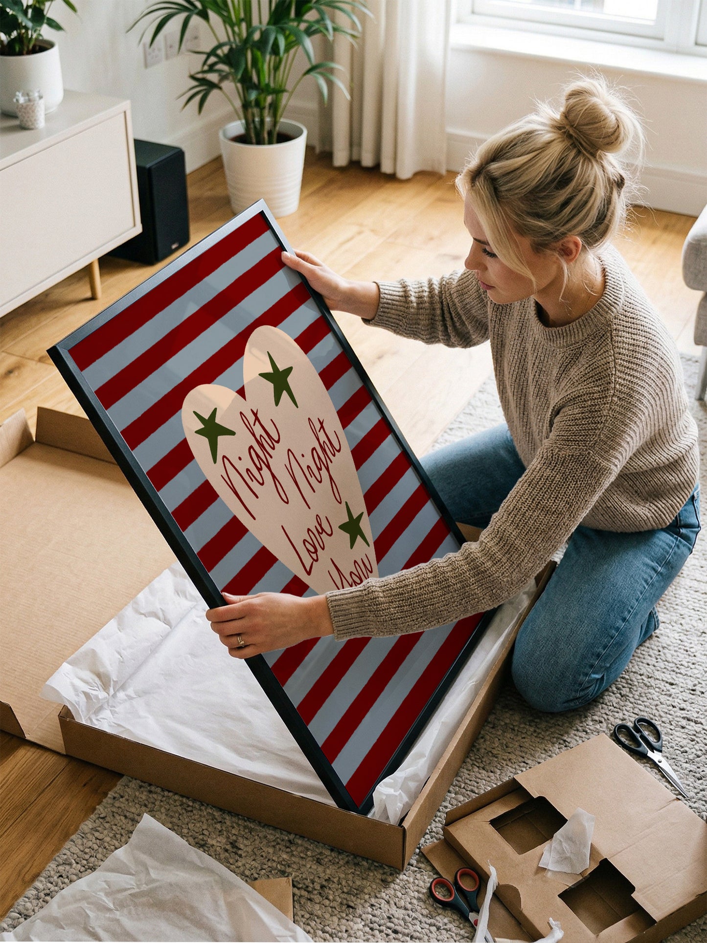A woman is kneeling on the floor and holding a large framed picture with a red and white striped background and a heart-shaped design.