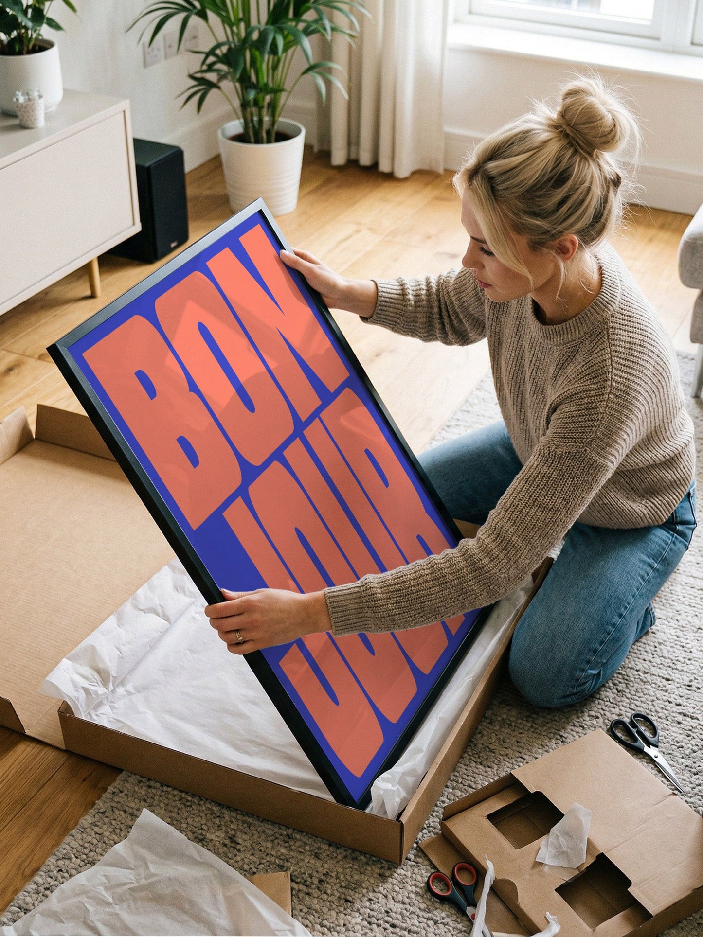 A woman is kneeling on the floor and holding a large framed poster with the word "BONJOUR" written on it.